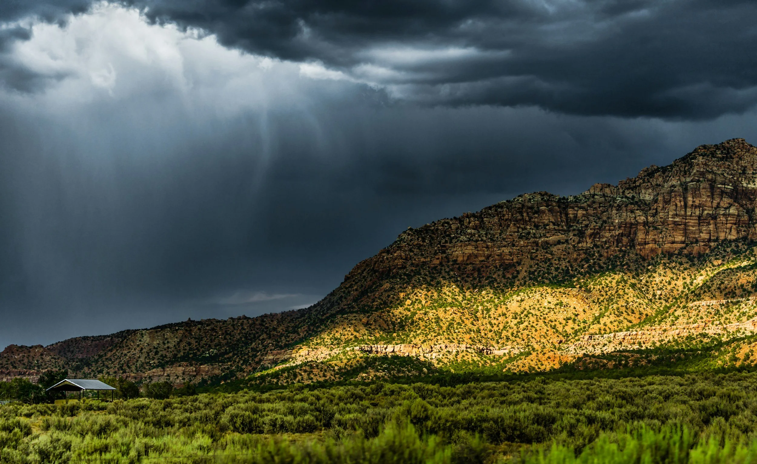 A mountain landscape under dark stormy clouds with rain in the distance, desert vegetation on the hills, and a small structure with a metal roof in the foreground.