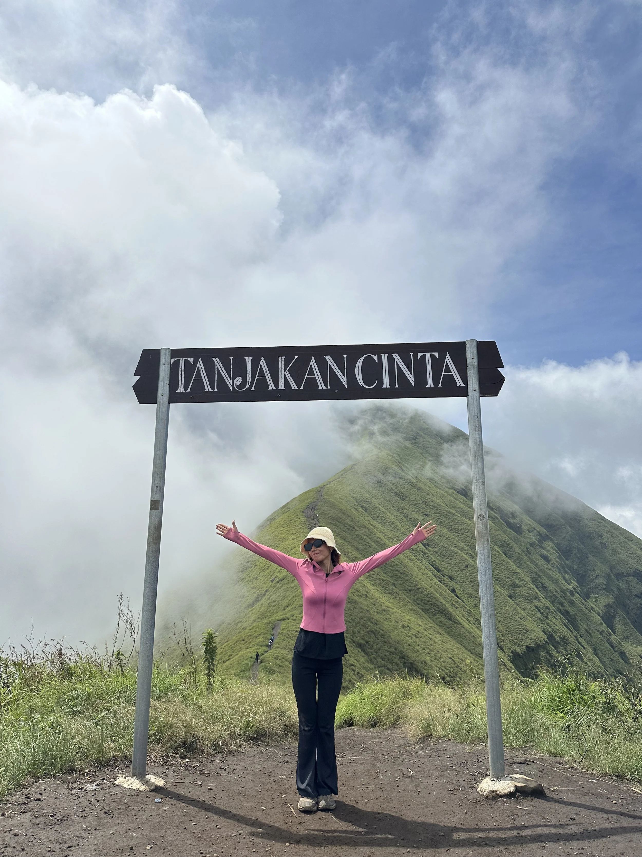 Woman in pink jacket and black pants standing with arms outstretched under a sign that reads 'Tanjakan Cinta' with a lush green mountain and a cloudy sky in the background.