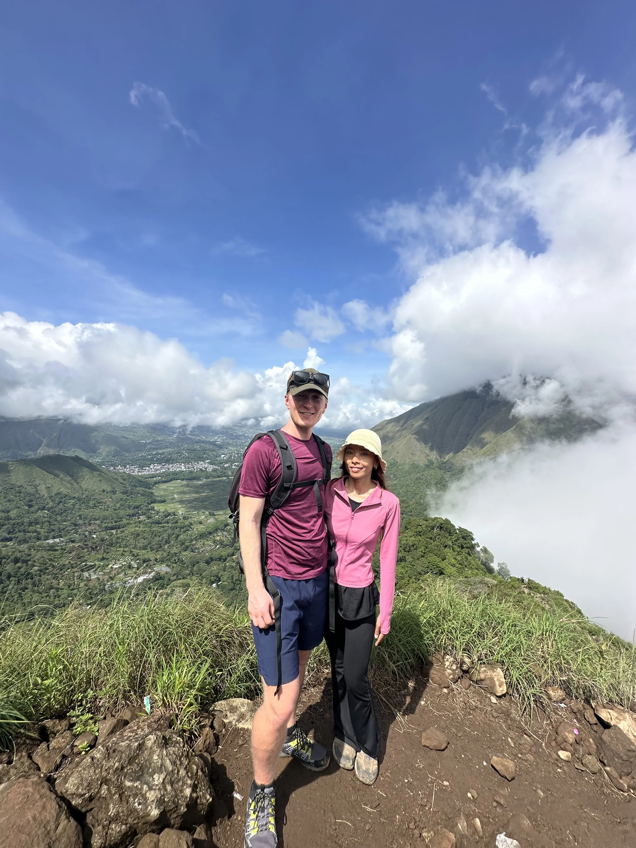 A man and woman smiling on a hiking trail with mountains, clouds, and a valley in the background.