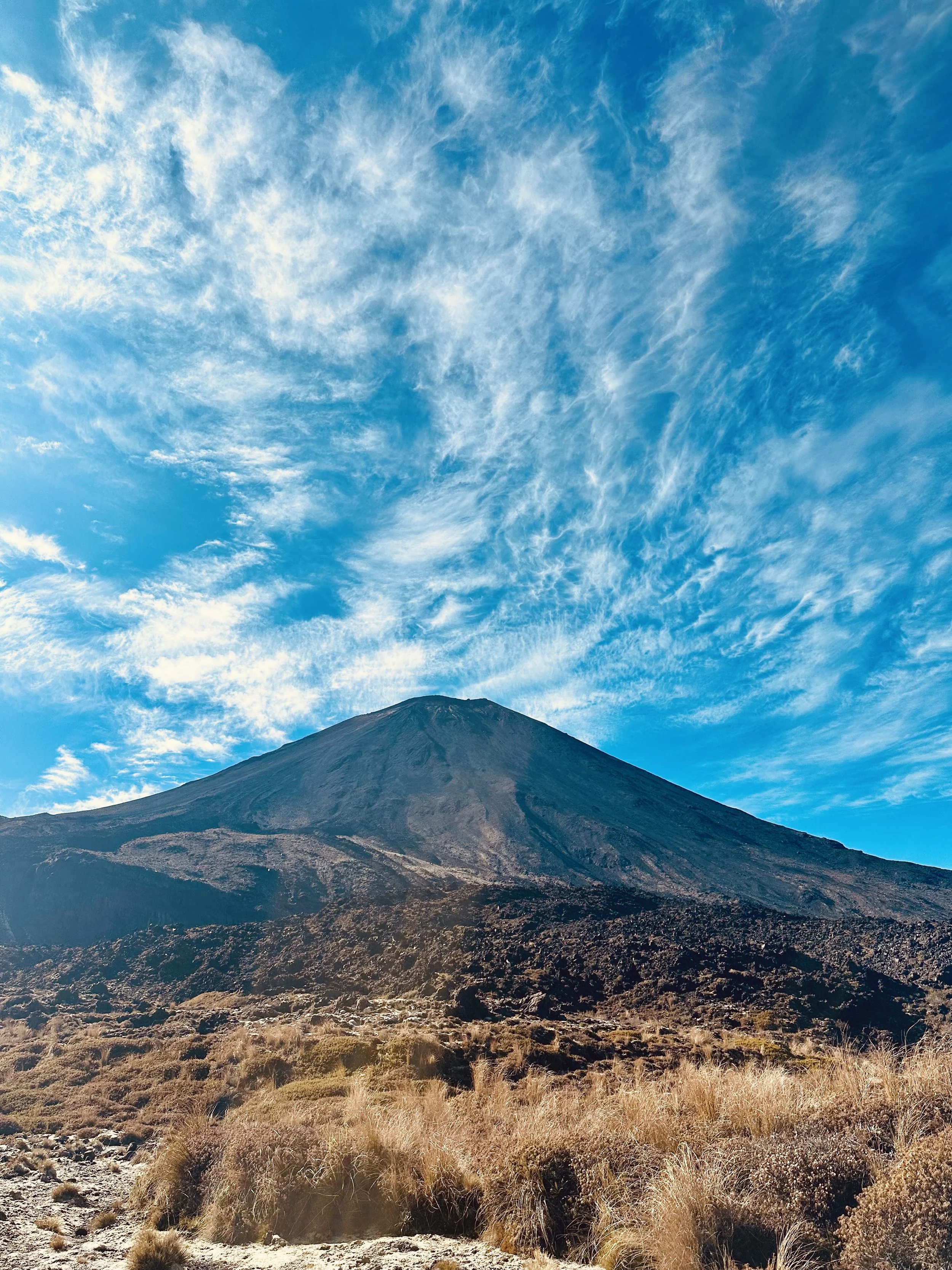 Volcano with a dark slope and rugged terrain in the foreground, under a bright blue sky with wispy clouds.