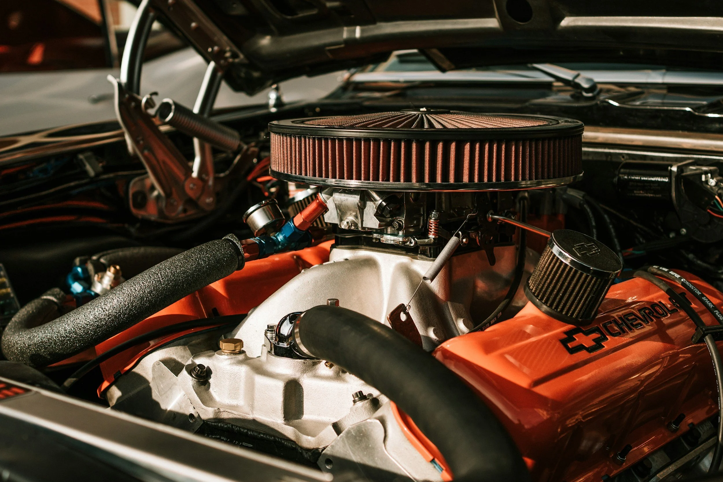 Close-up of a car engine with a round air filter on top, various hoses and components visible.