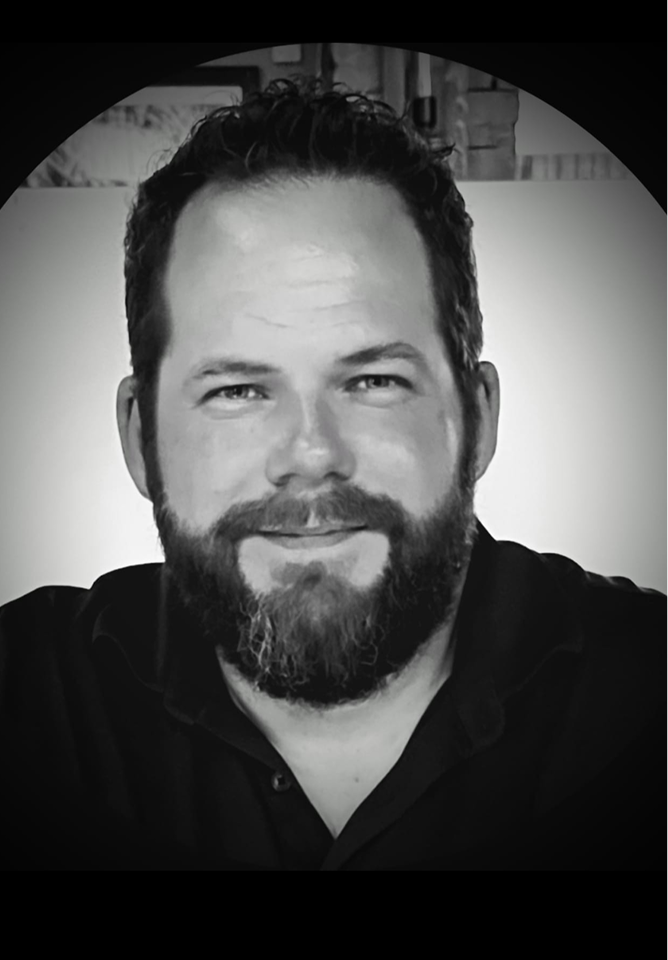 Black and white photo of a smiling man with a beard and short hair, wearing a dark shirt, sitting indoors, with a blurred background and an unusual circular framing.