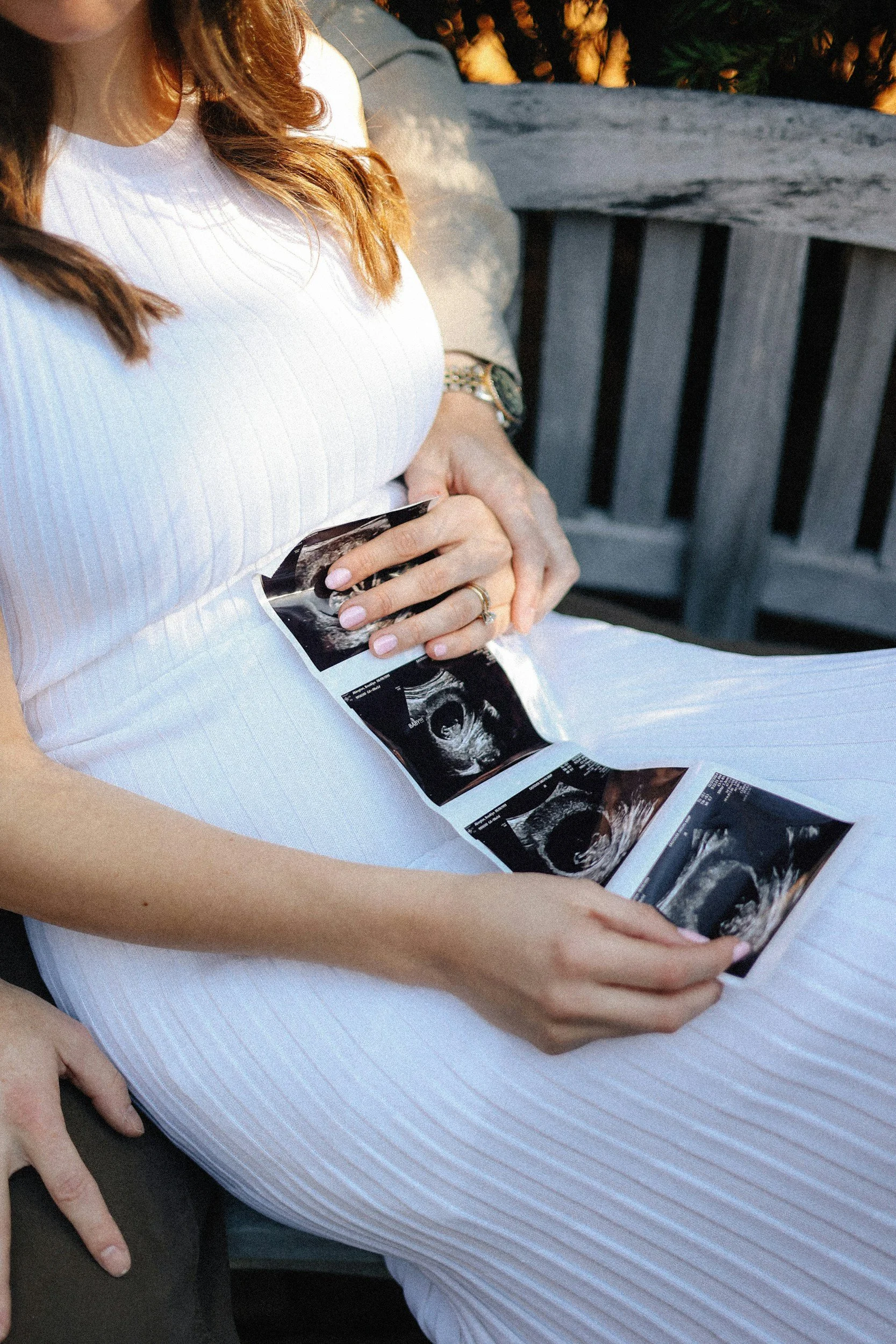 A pregnant woman with red hair, wearing a white dress, sitting outdoors on a bench, holding ultrasound images of her unborn baby.