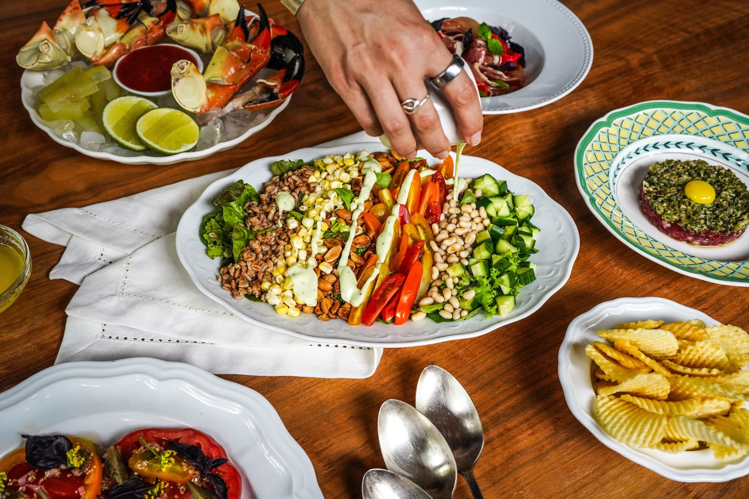 A person is serving salad from a large white platter containing mixed vegetables like cherry tomatoes, cucumbers, and beans, with dressing being poured on top. Surrounding this are various dishes including a seafood platter with lime wedges, a dish with an egg on top, and a bowl of ridged potato chips.