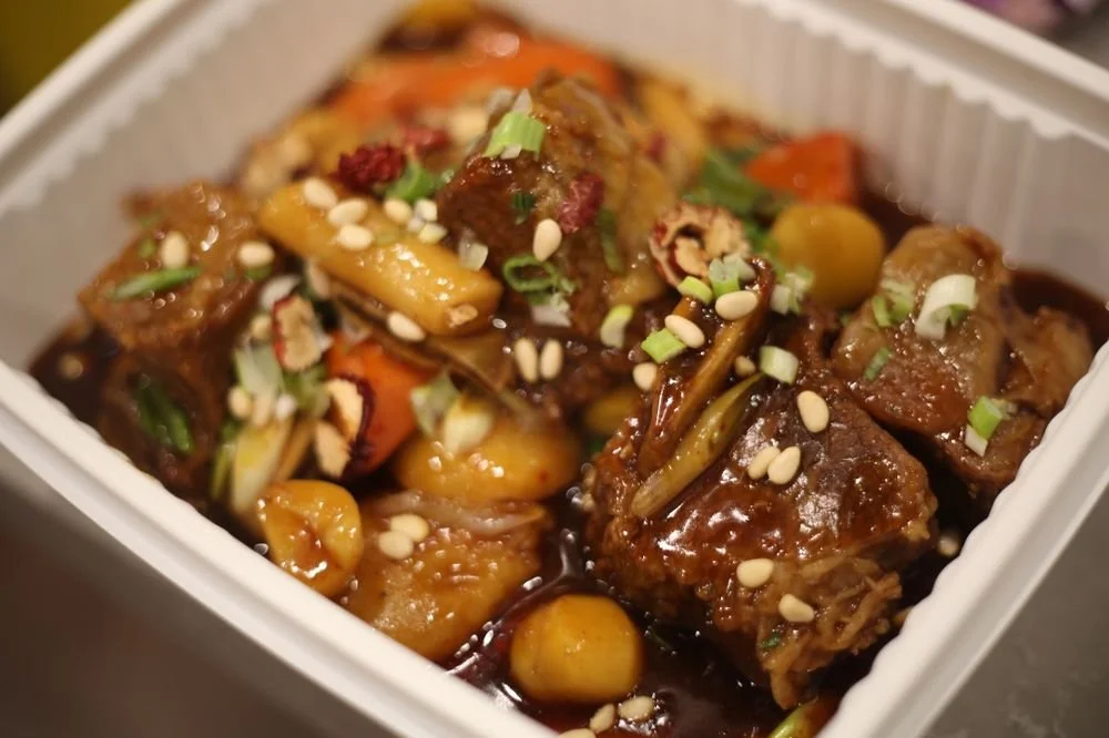 Close-up of Chinese takeout dish with beef, vegetables, and sauce topped with chopped green onions and sesame seeds.