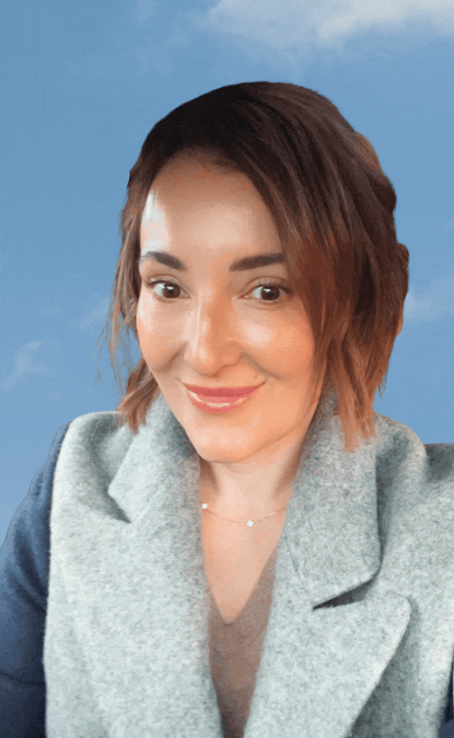A woman with short, wavy brown hair and light makeup, smiling against a blue sky background.