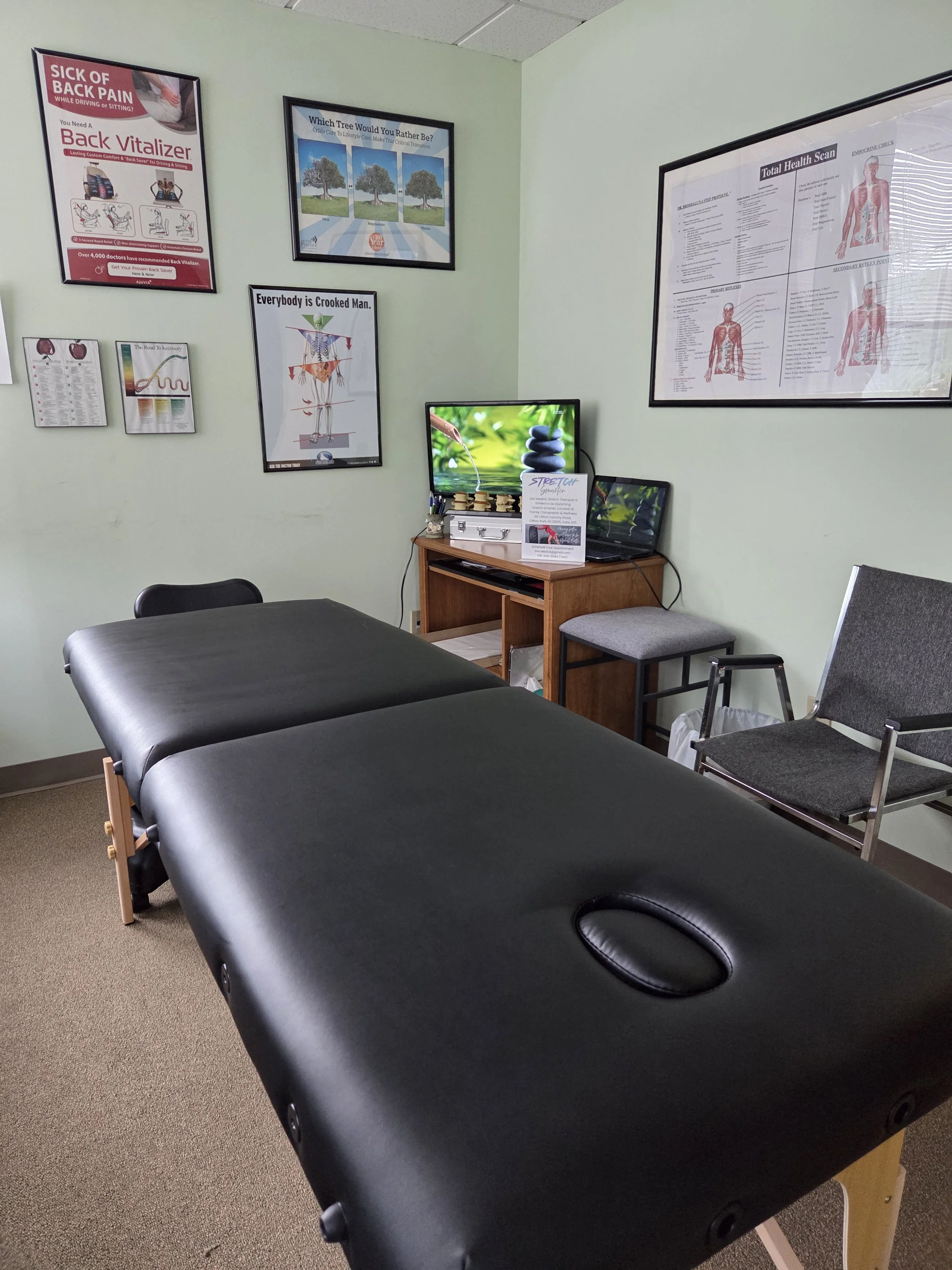 A therapy or massage room with a black massage table, a black chair, and a small wooden cabinet with a computer monitor showing a stack of stones and a pamphlet about stretching. The wall has posters, including one about back pain, a diagram of a human body, and wellness information.
