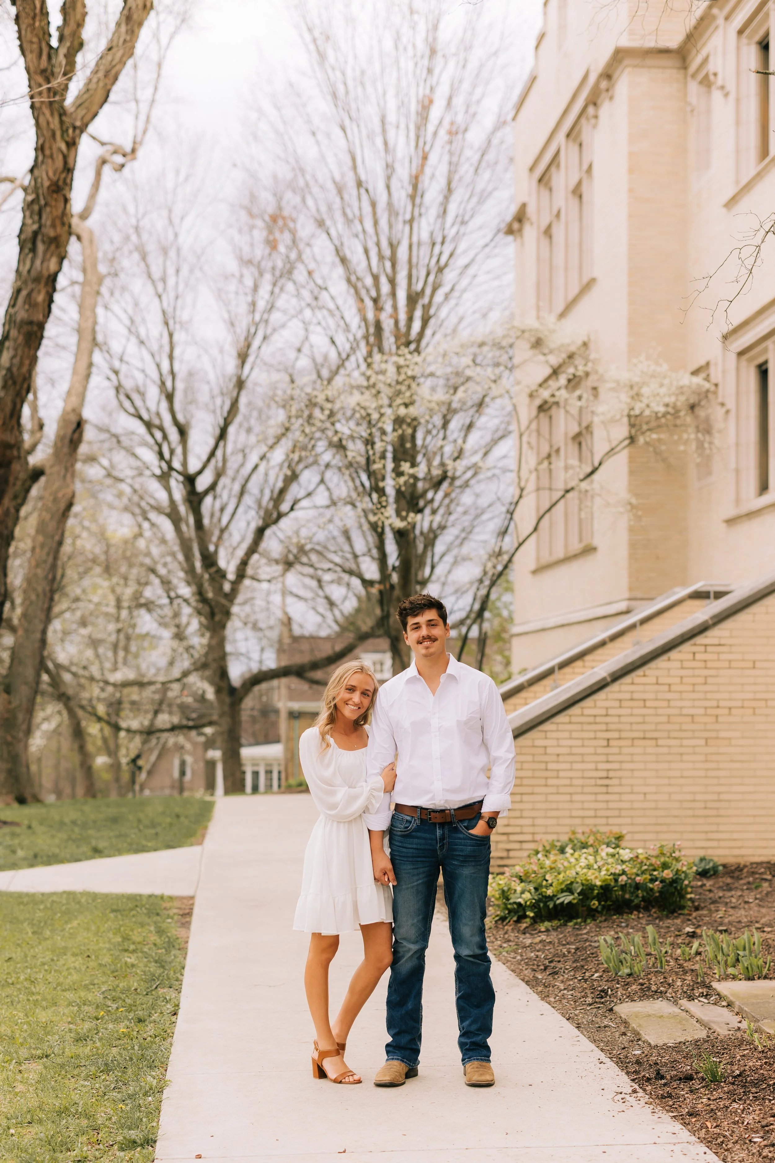 A couple posing outdoors on a sidewalk in front of a brick building with trees in spring bloom.