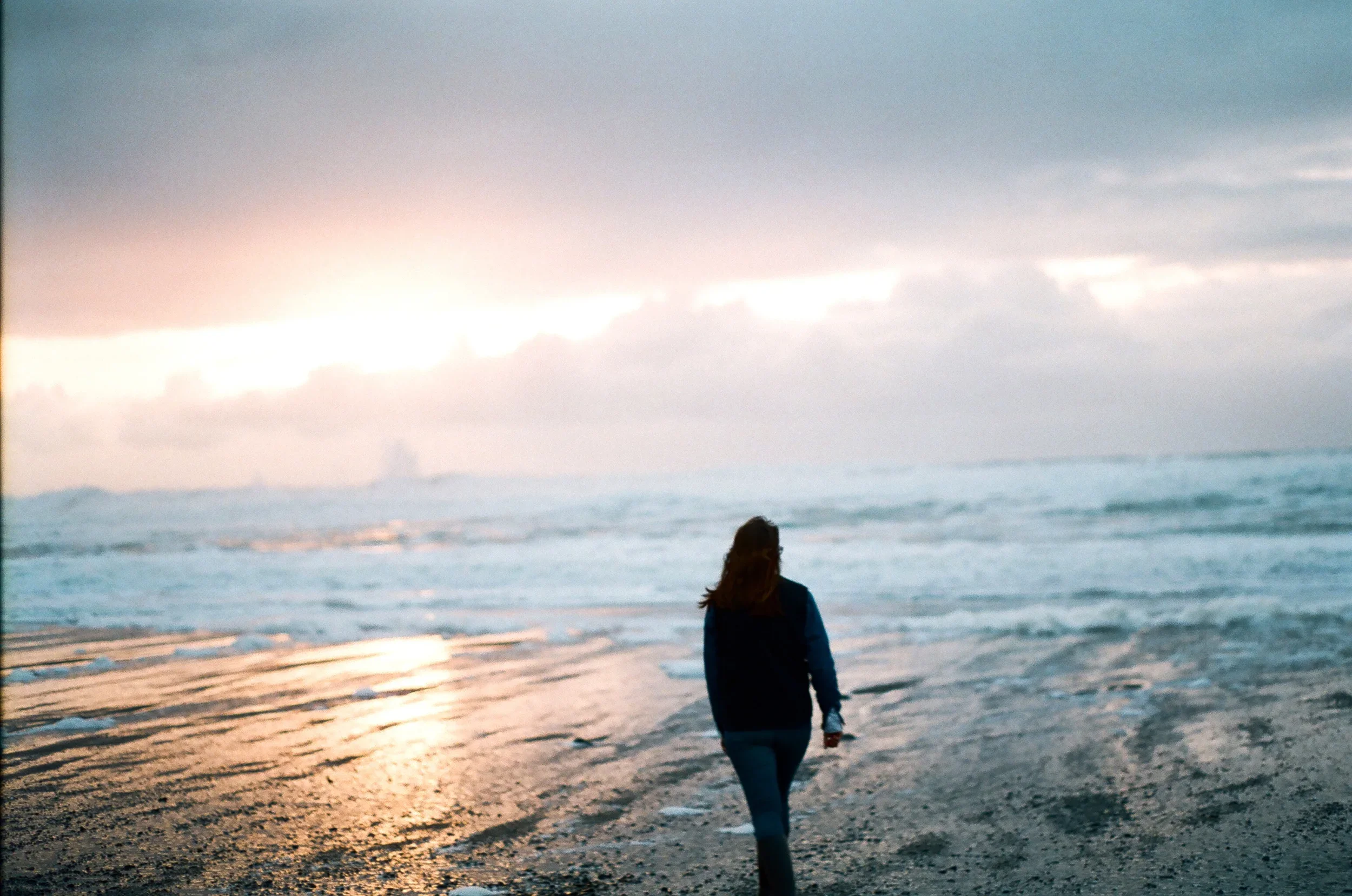 A person with long hair walking on the beach at sunset, wearing a dark jacket, with the ocean and cloudy sky in the background.