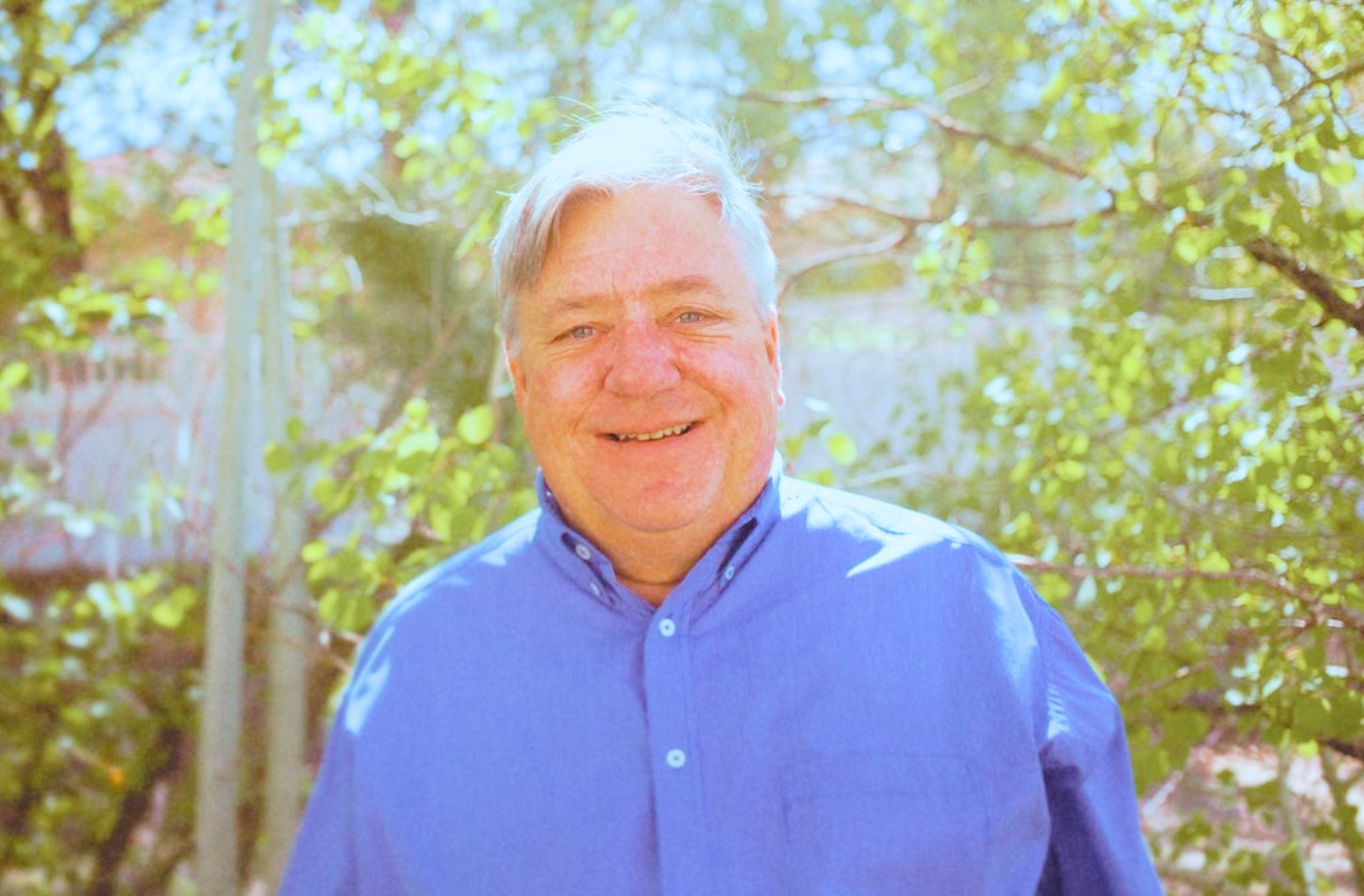 Smiling man with gray hair in a blue shirt outdoors surrounded by green foliage on a sunny day.