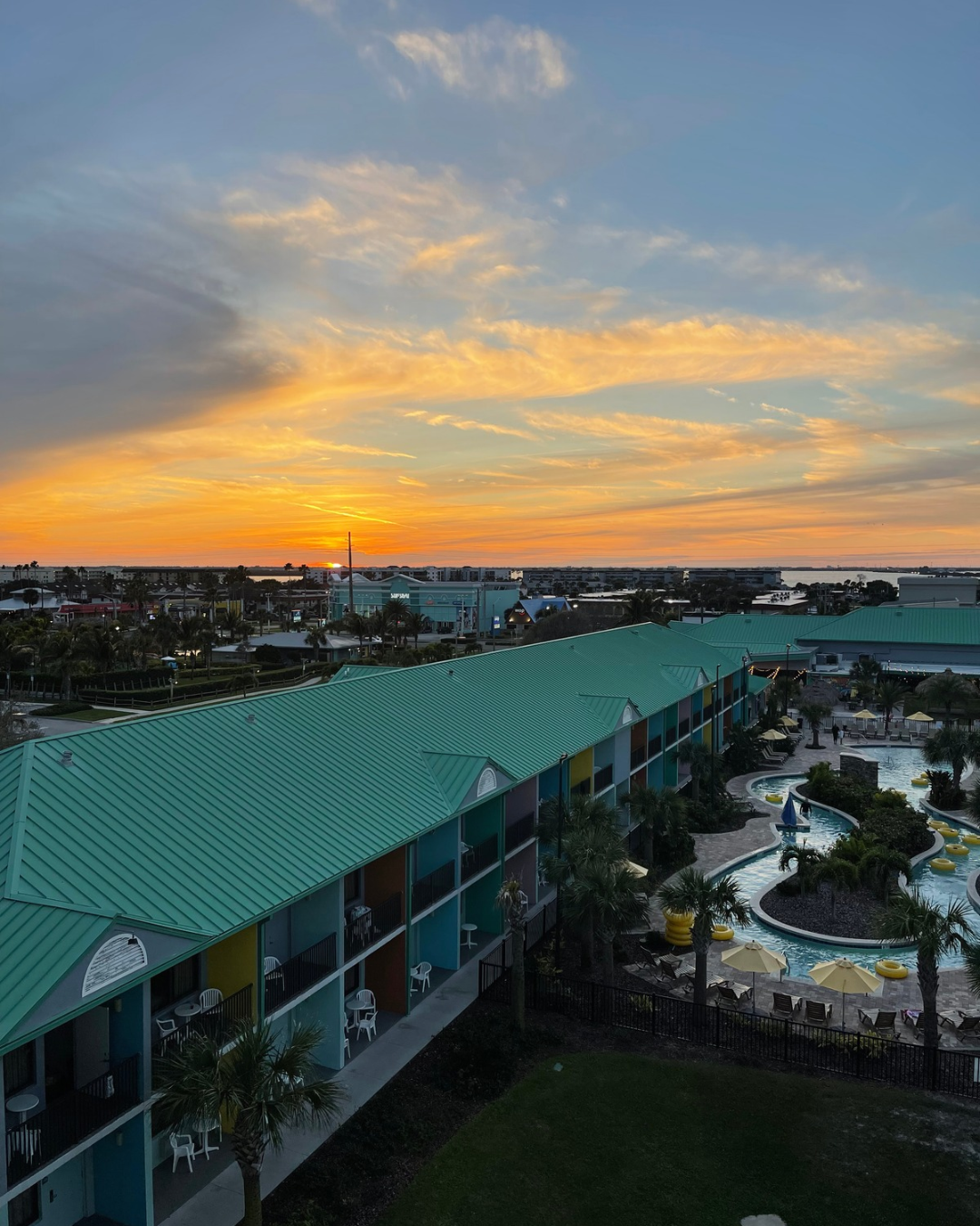Sunset over a colorful hotel with teal rooftops, outdoor pool with yellow floaties, palm trees, and a clear sky with orange and blue clouds.