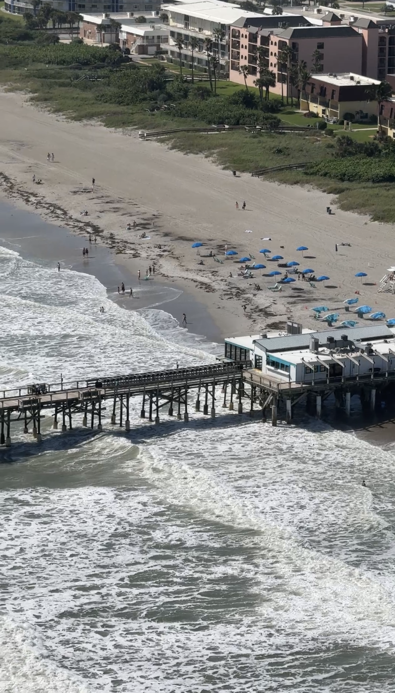 A beach scene with people walking and relaxing under blue umbrellas, along with a pier extending into the ocean and residential buildings in the background.