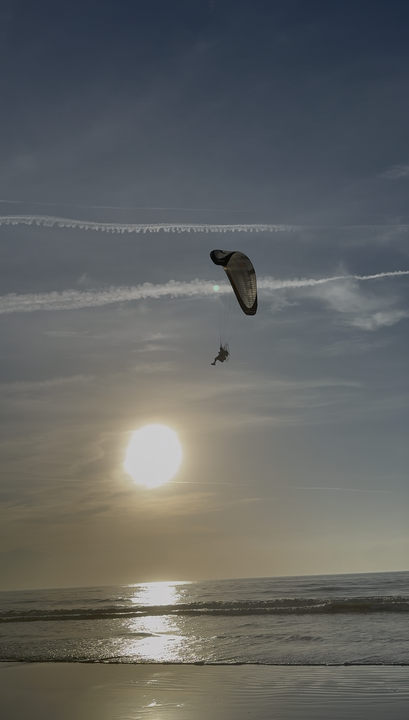 A person paragliding over the ocean at sunset, with the sun near the horizon and waves along the shoreline.