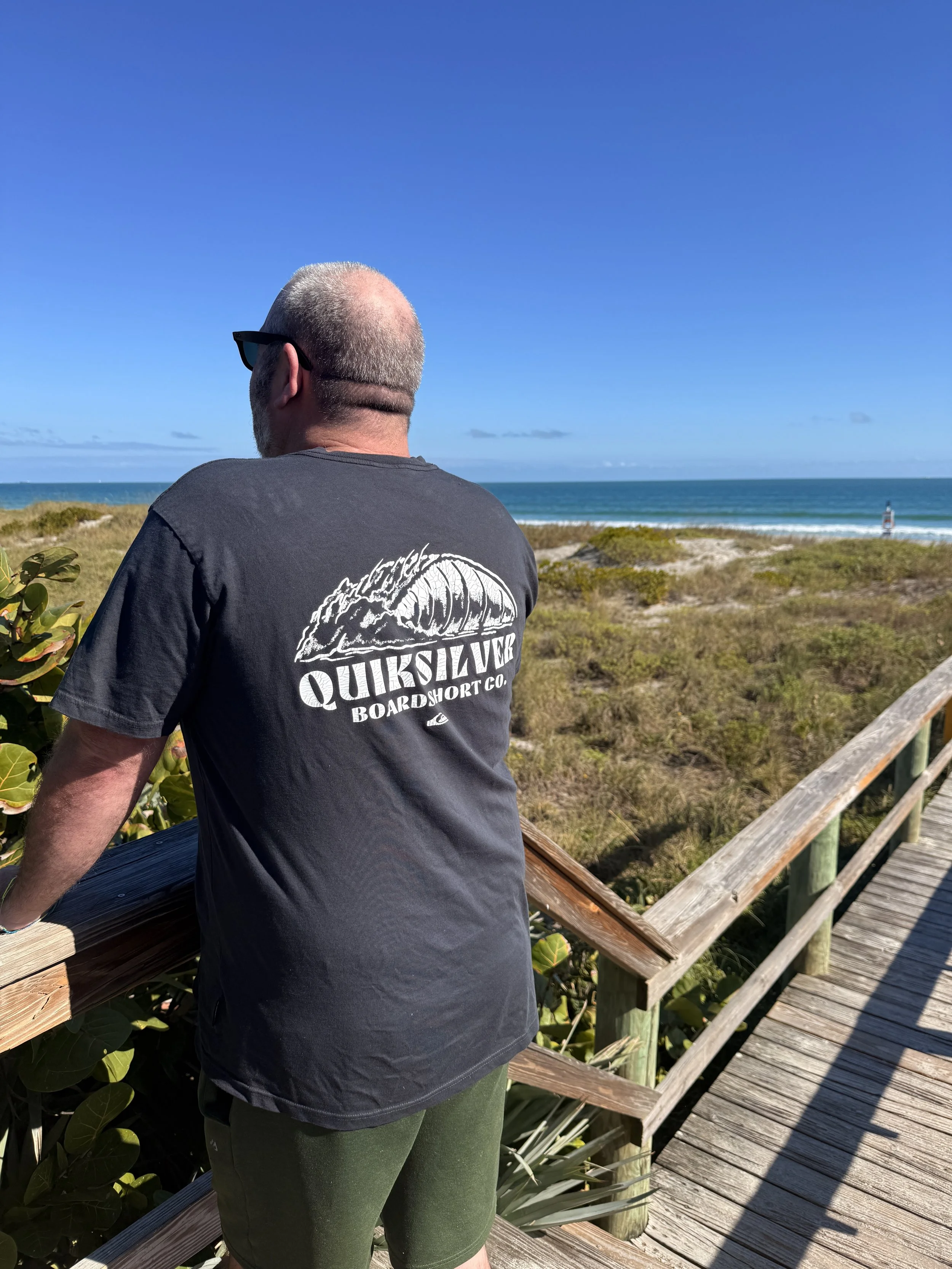 A man wearing glasses, a Quiksilver t-shirt, and green shorts standing on a wooden boardwalk overlooking a beach with blue sky and ocean in the background.