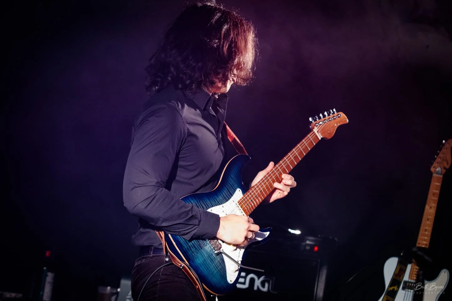 A person with long, curly hair wearing a dark shirt playing an electric guitar on stage, with dark background and stage lighting.
