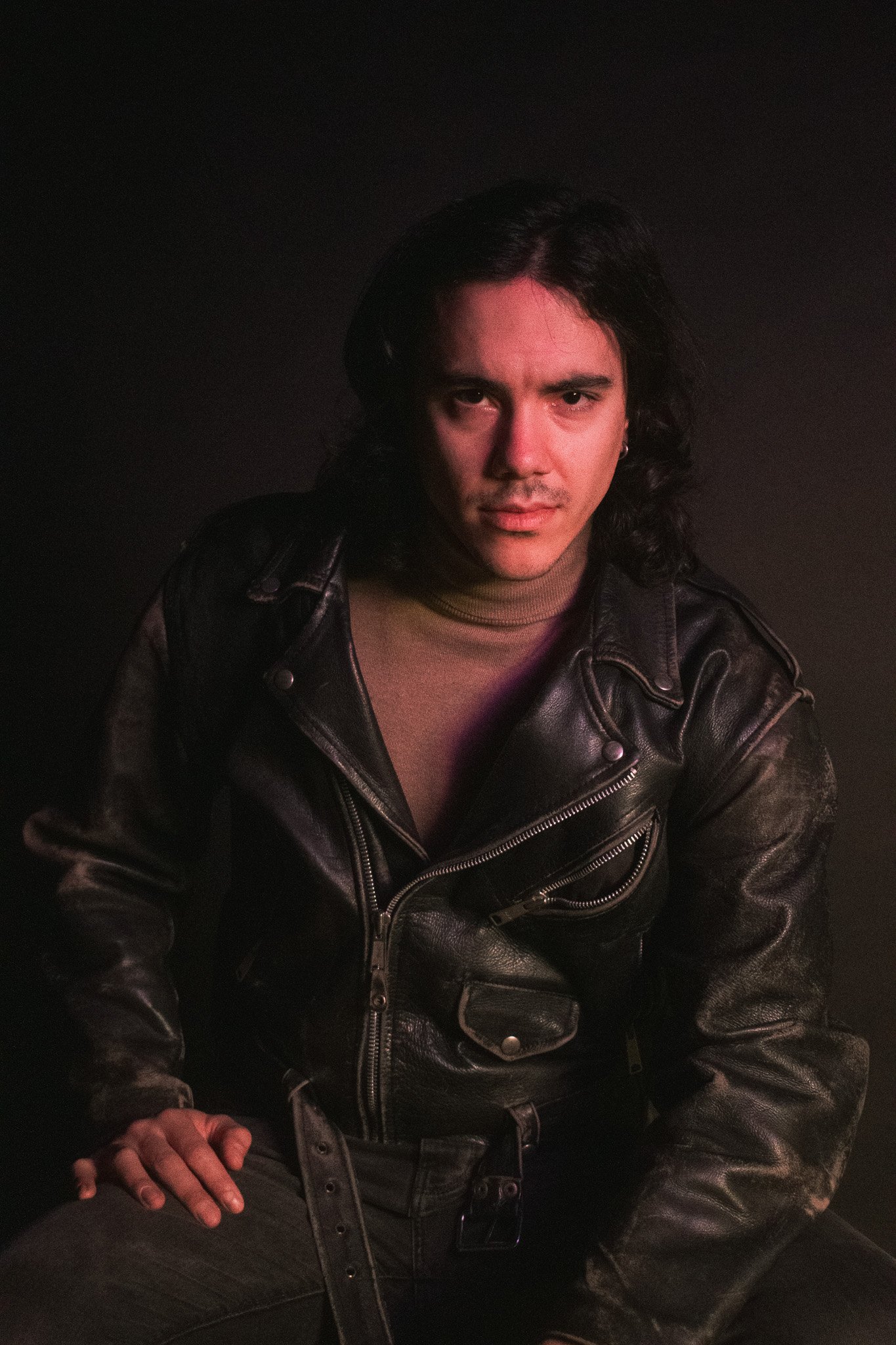 A man with shoulder-length dark hair and light skin wearing a black leather jacket, tan turtleneck, and black pants, sitting in low lighting.