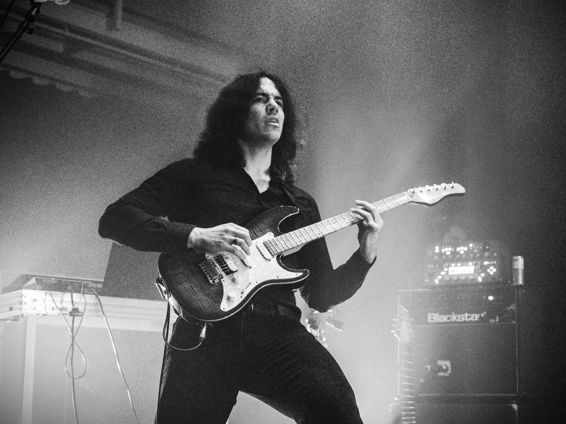 A person with long dark hair playing an electric guitar on stage, with equipment and monitors visible in the background.