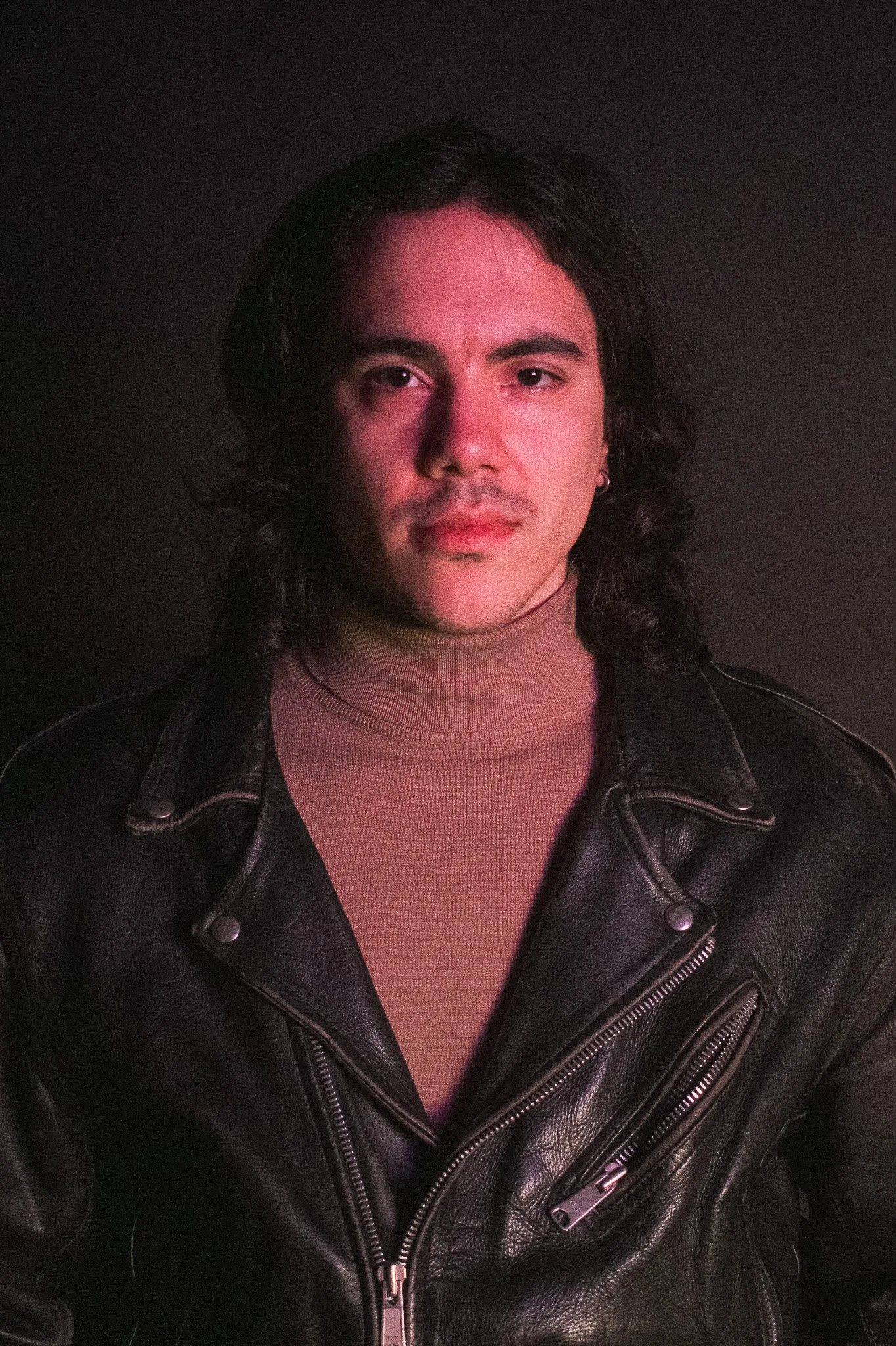 A person with long dark hair, wearing a beige turtleneck and black leather jacket, standing against a dark background.