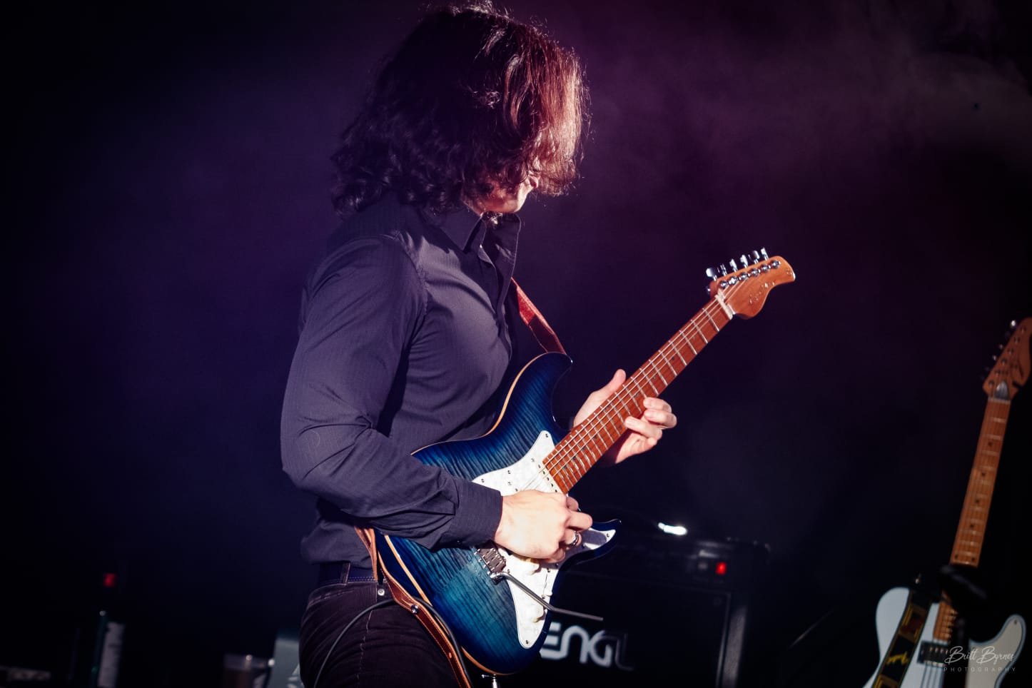 A person with curly hair playing an electric guitar on stage with a dark background.