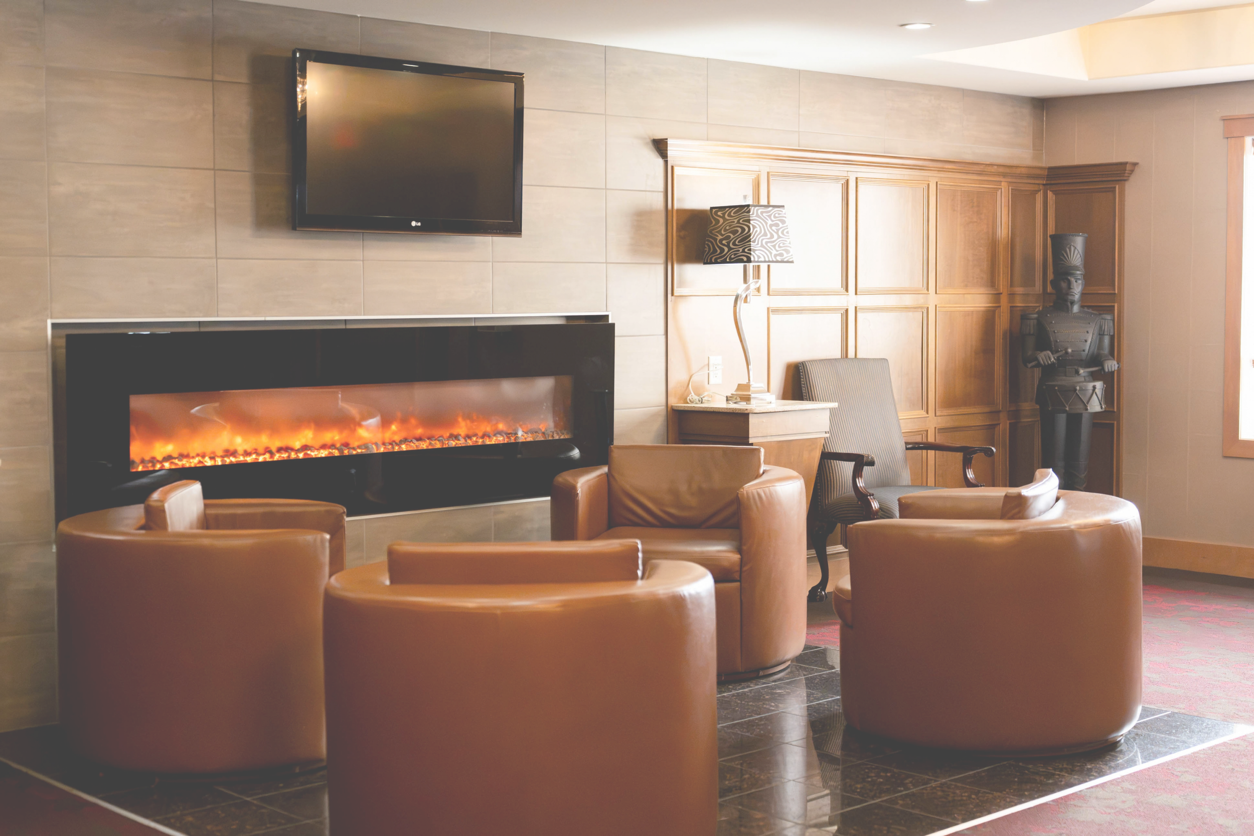 A hotel-style lobby area with leather armchairs facing a modern electric fireplace, a wall-mounted flat-screen TV, a wooden cabinet, and a tall decorative statue of an ancient warrior.