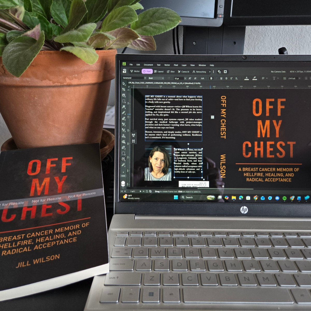A potted plant with green leaves on a desk next to a computer screen and a paperback book titled "Off My Chest" by Jill Wilson, with the subtitle "A Breast Cancer Memoir of Hellfire, Healing, and Radical Acceptance." The computer screen displays a book cover design and some text for the same book.