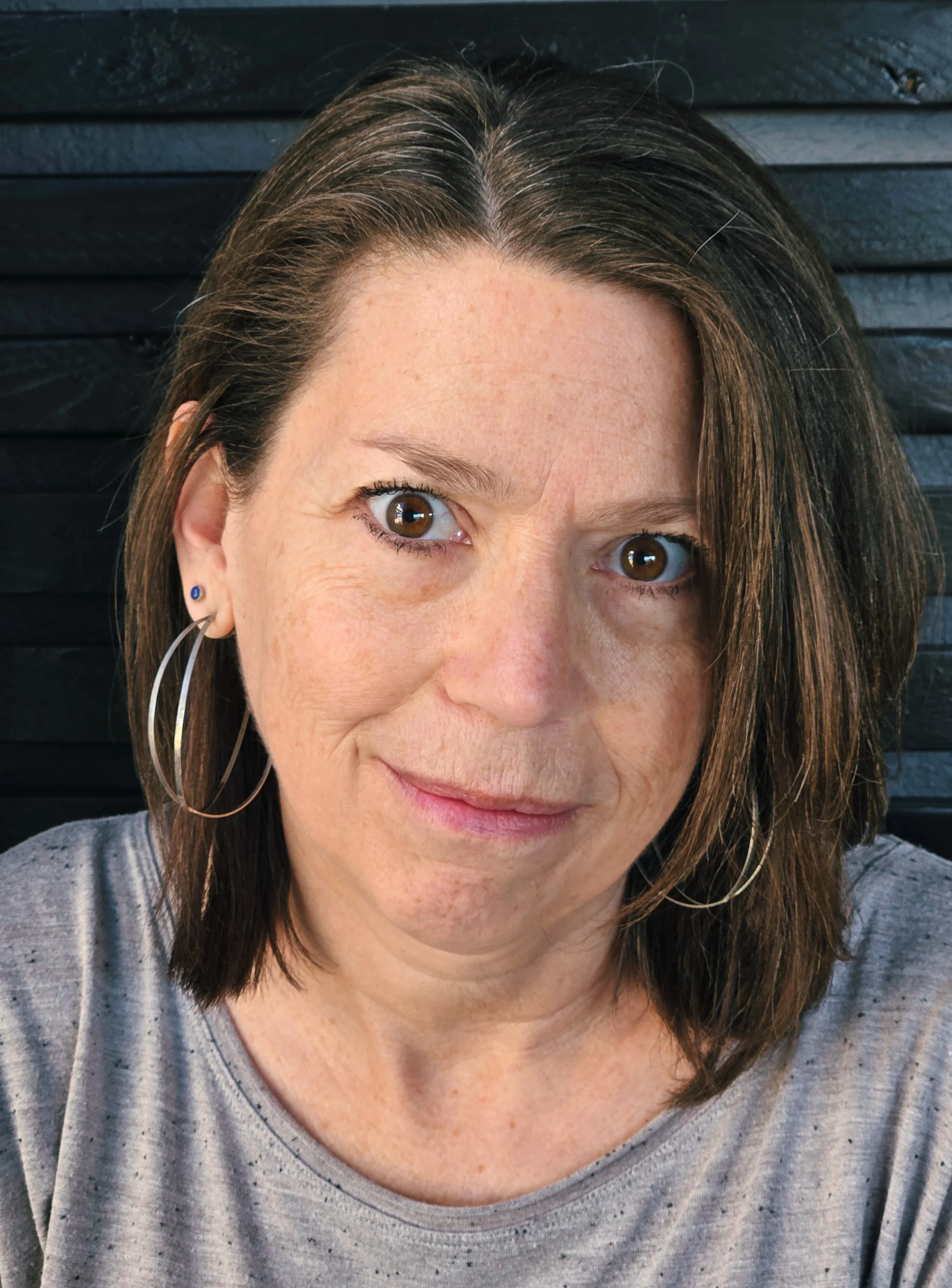 A woman with brown hair and brown eyes wearing a grey shirt, looking at the camera, with a black wall as background.