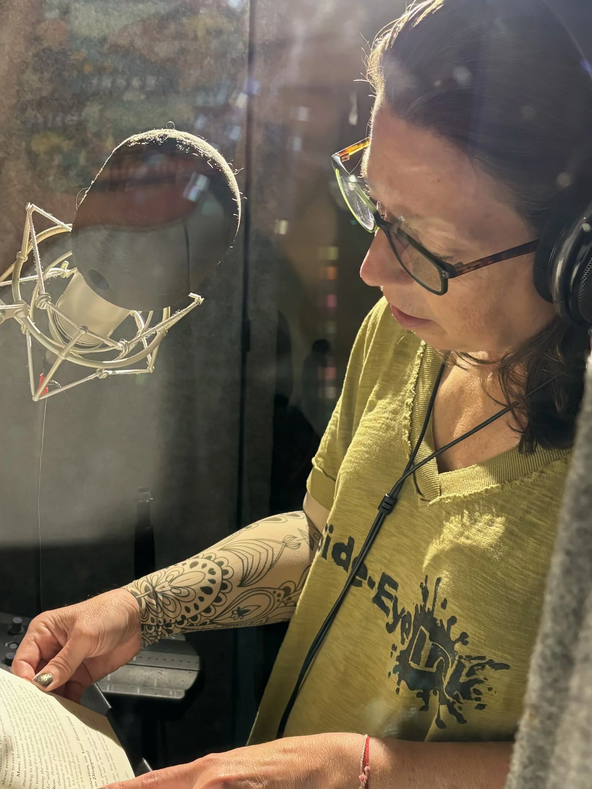 A woman with dark hair, glasses, and a yellow t-shirt is sitting in a recording booth, reading a book with a microphone in front of her. She has tattoos on her arms and is wearing headphones.
