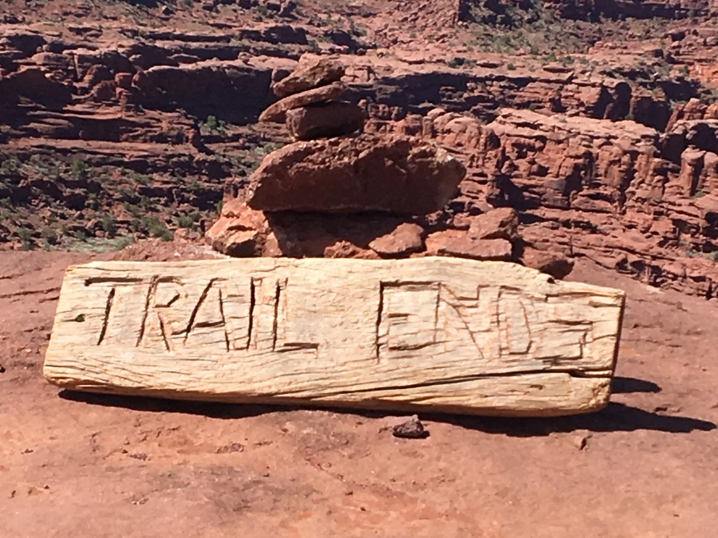 A wooden sign with the wordS 'TRAIL ENDS' carved into it, leaning against a small pile of balanced rocks in a desert landscape with reddish rocks and sparse vegetation.