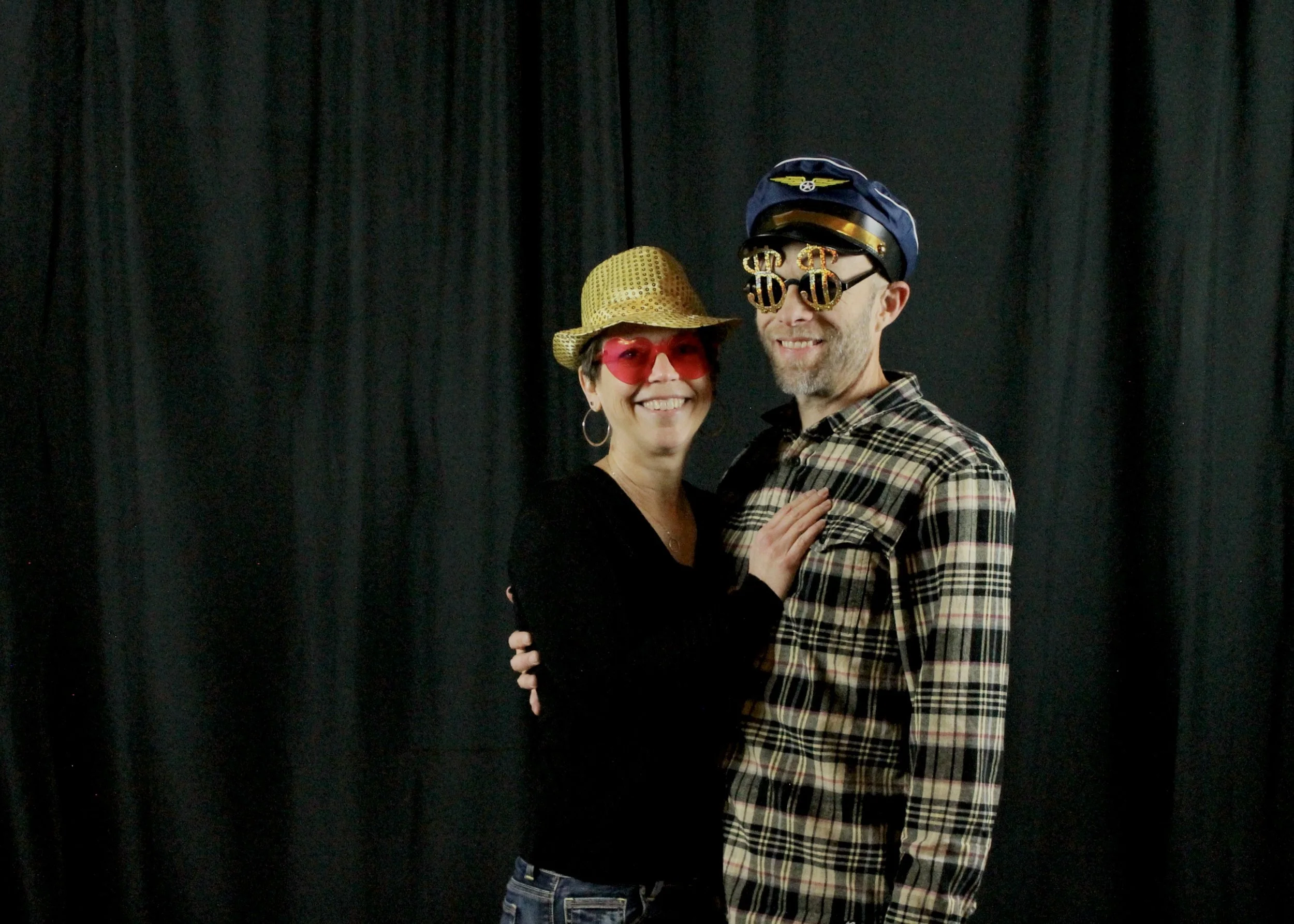 A smiling woman and man posing together against a black curtain. The woman is wearing red sunglasses and a gold hat, while the man is wearing glasses with dollar sign decorations and a pilot cap, both dressed casually.