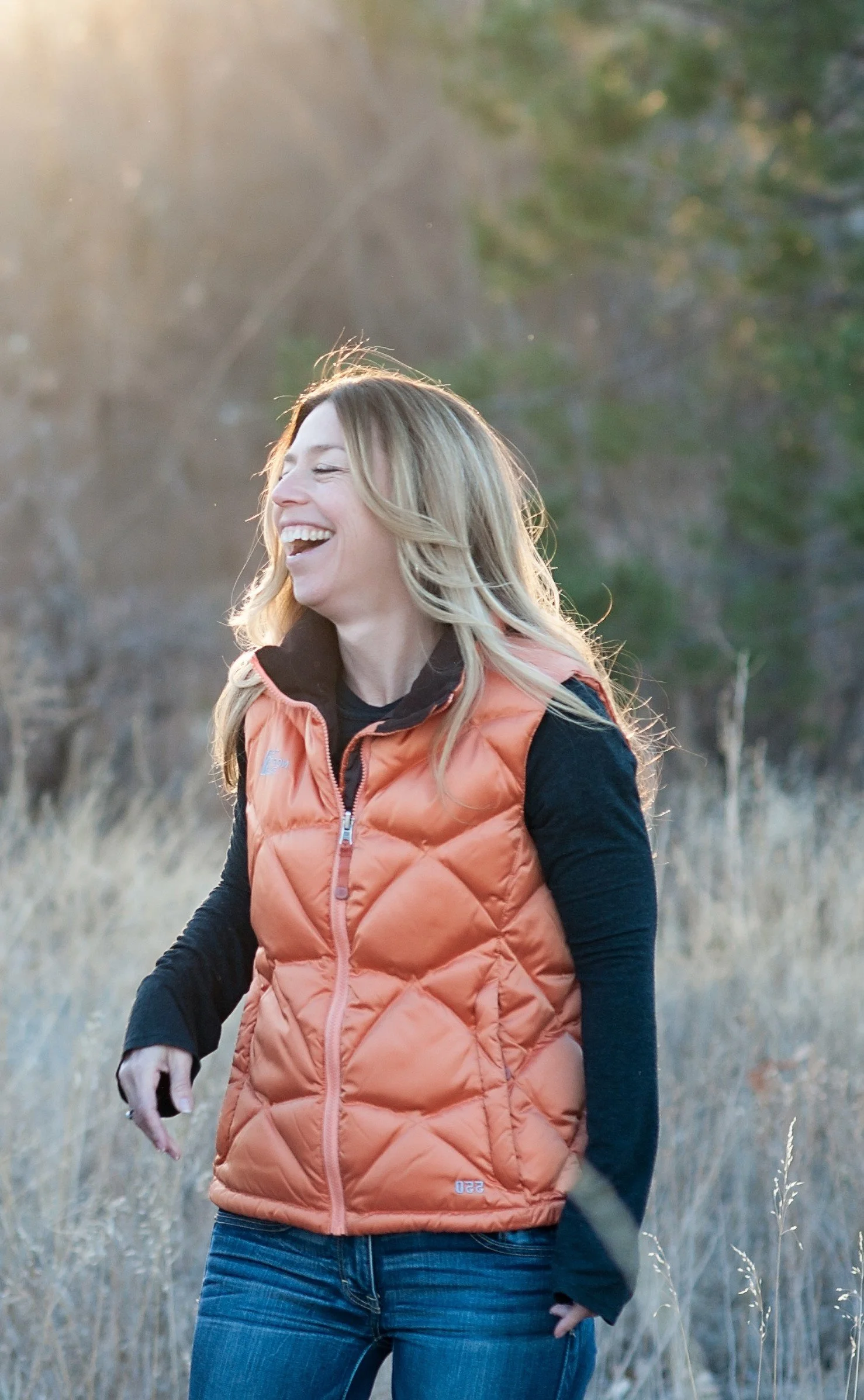 A woman with blonde hair smiling outdoors during sunset, wearing an orange quilted vest and black long-sleeve shirt, standing in a field with trees in the background.