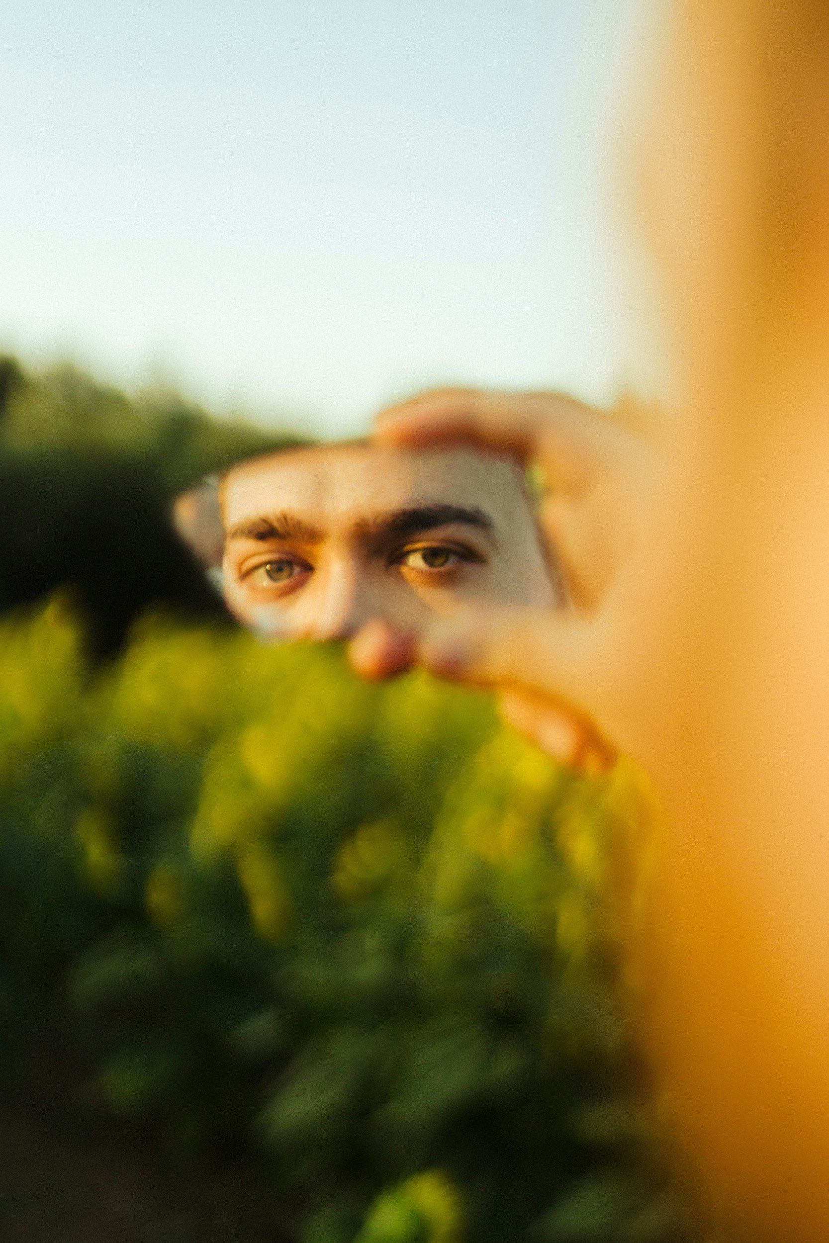 Mann steht in einem Feld und hält einen kleinen Spiegel vor sein Gesicht, in dem seine Augenpartie zu sehen ist.
