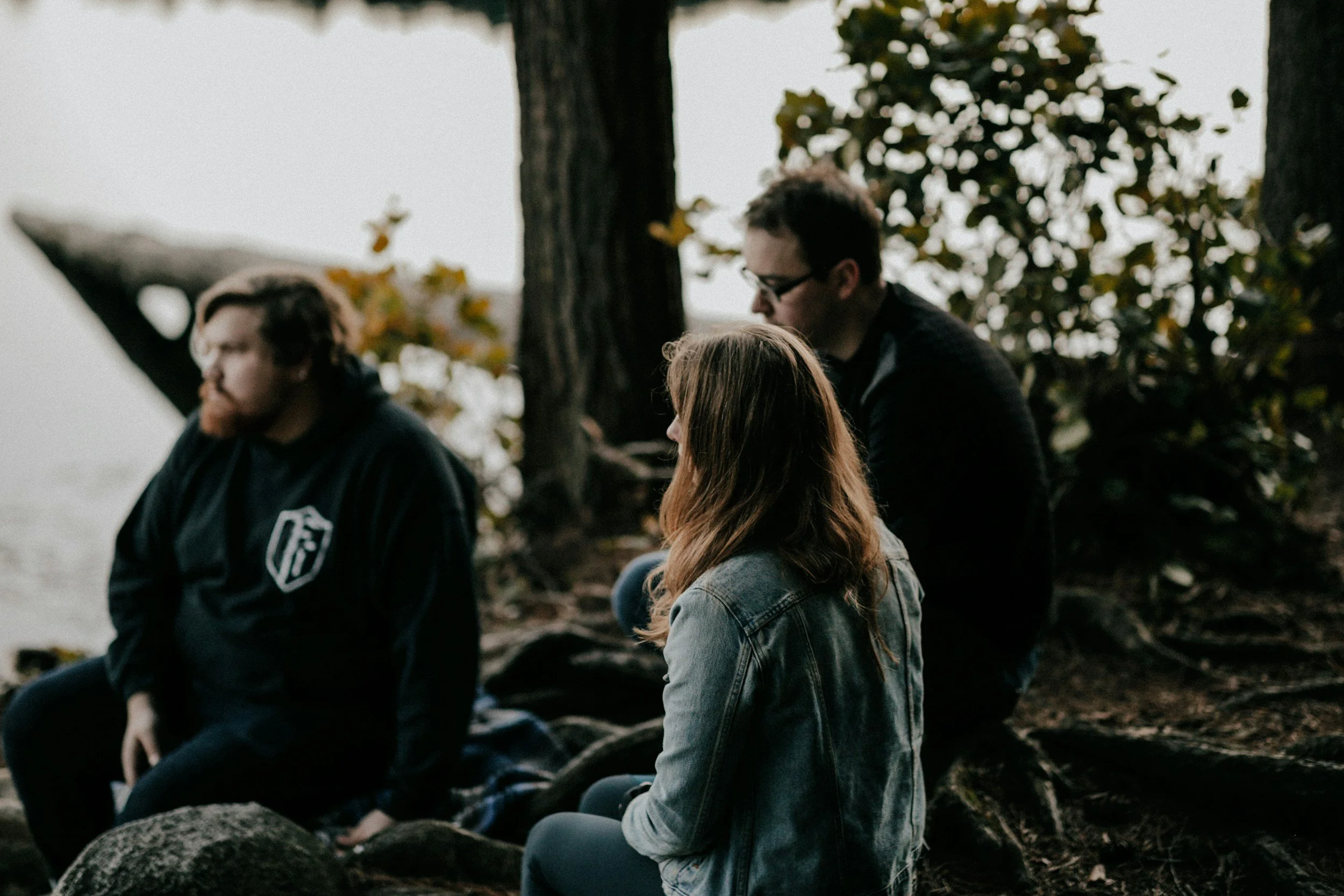 Drei Menschen sitzen im Wald auf Steinen, eine Person hat rote Haare, eine trägt eine Jeansjacke, alle scheinen nachdenklich oder meditativ.