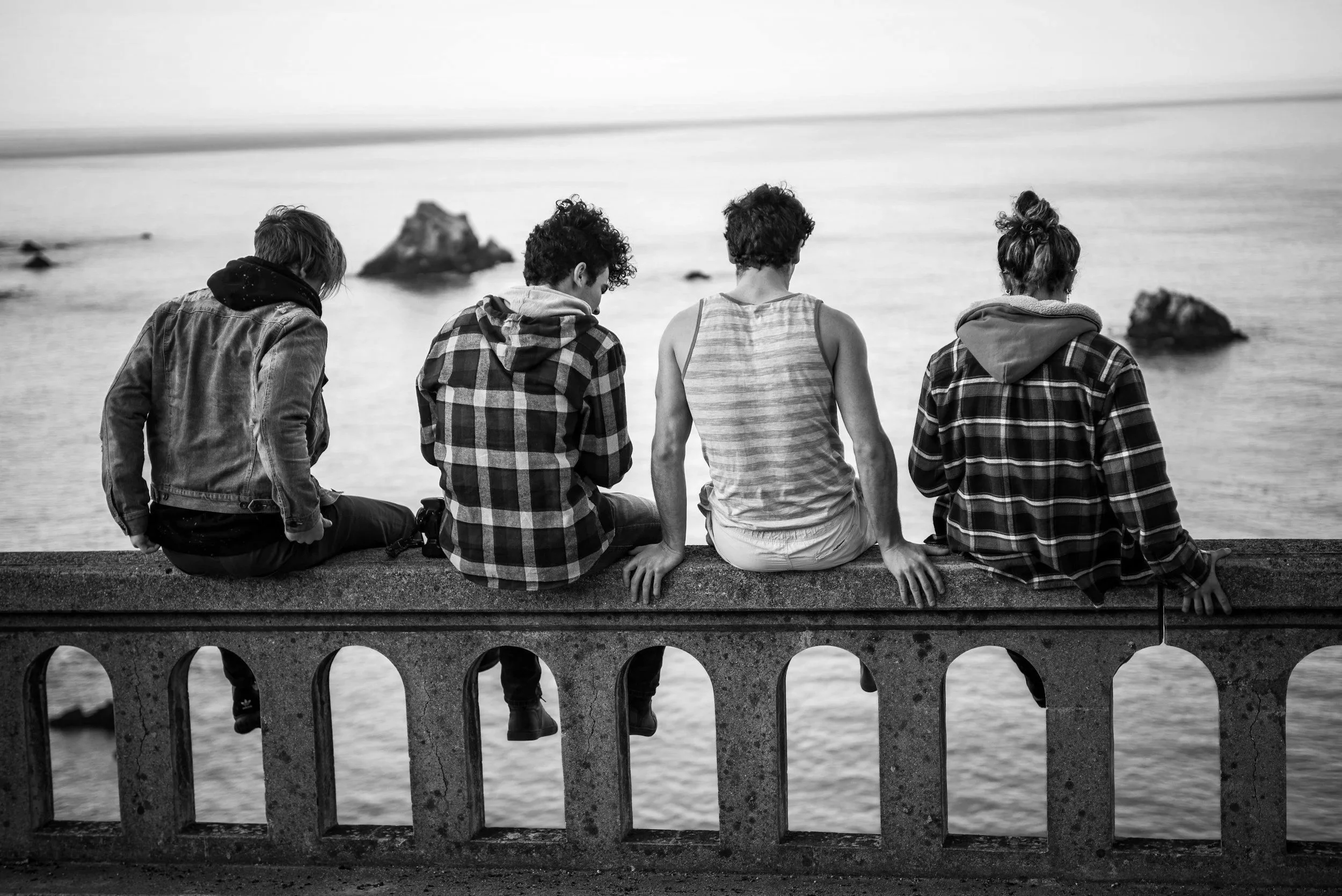 Vier Jugendliche sitzen auf einer Brüstung am Meer, blicken ins Wasser, tragen Casual Kleidung, bei Tageslicht, Schwarz-Weiß-Foto.
