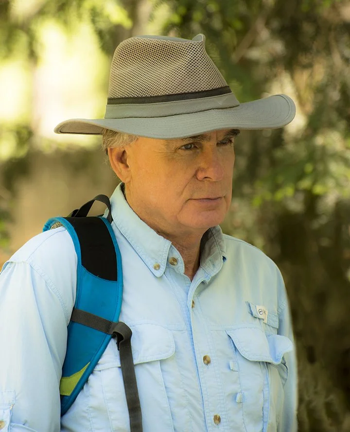 An older man wearing a wide-brimmed hat, a light blue button-up shirt, and a backpack, standing outdoors with a blurred green background.