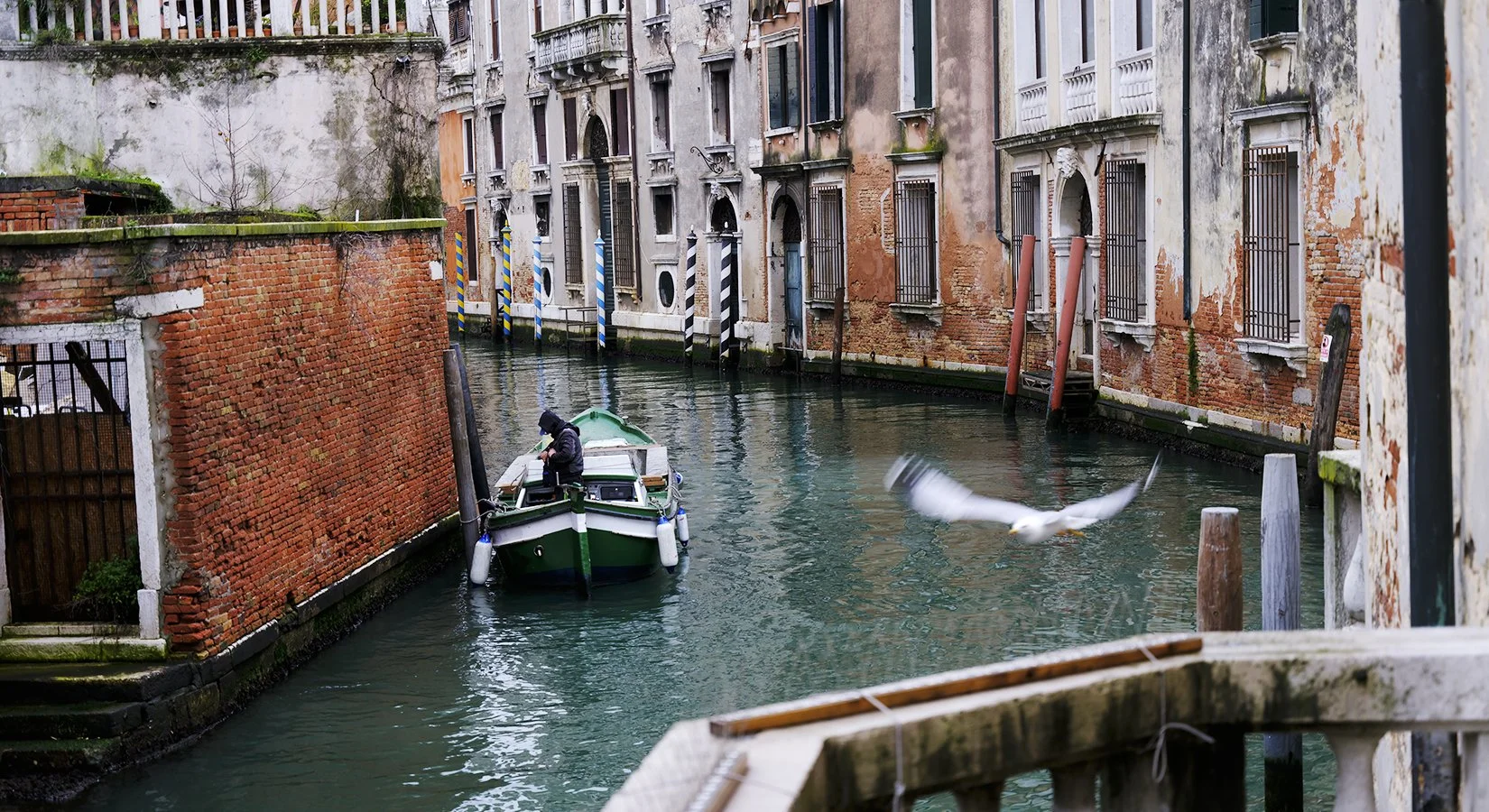 A person in a black coat and hood stands on a small boat in a narrow canal with old, weathered buildings with barred windows on either side. A seagull flies nearby.