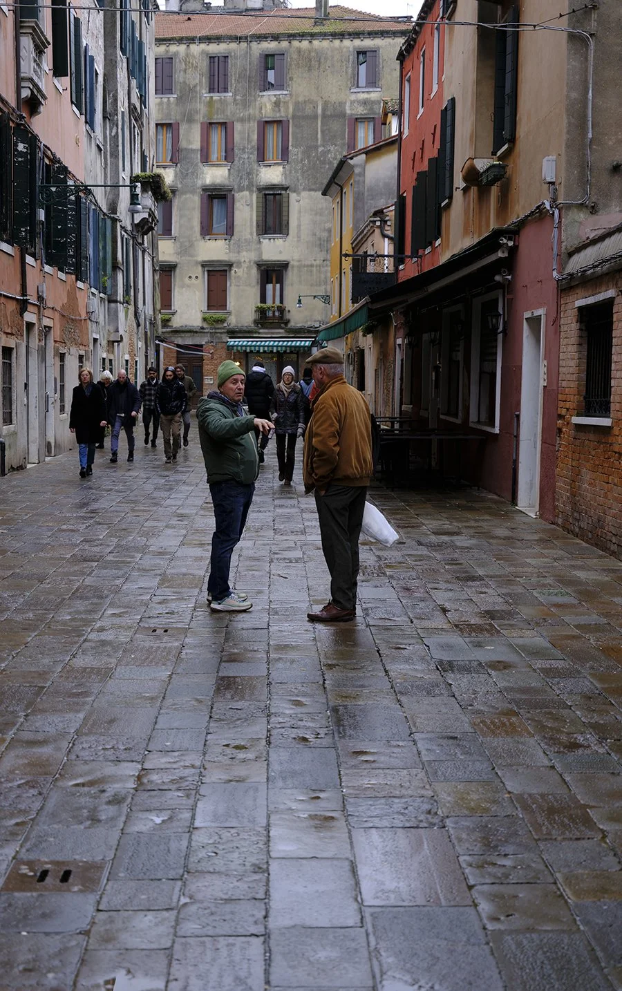 Two men are talking on a wet cobblestone street in a European city with colorful buildings on either side and pedestrians walking in the background.