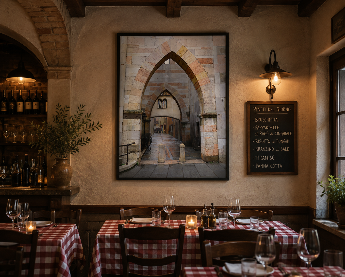 Interior of a cozy Italian restaurant with tables covered in red and white checkered tablecloths, wine glasses, lit candles, and a chalkboard menu on the wall listing daily specials. There is a large framed photograph of an ancient stone archway street scene and greenery in vases.