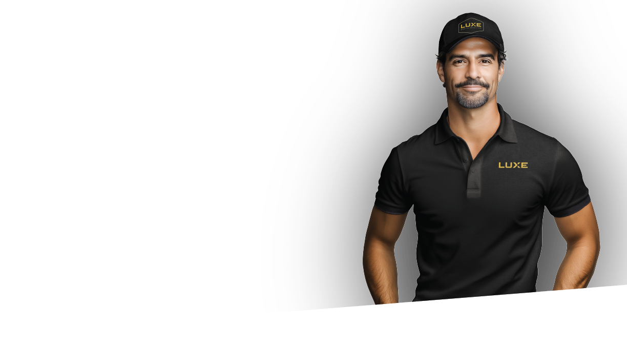 A man in a black polo shirt and black cap with the logo "LUXE" stands against a black background with a white angled bottom edge. The man is smiling subtly, with a short beard and mustache.