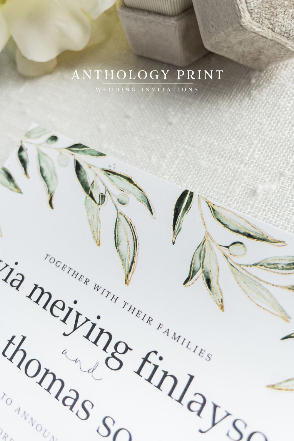 Close-up of a wedding invitation card with green, gold-accented leaf illustrations, placed on textured fabric with decorative boxes and a white flower in the background.