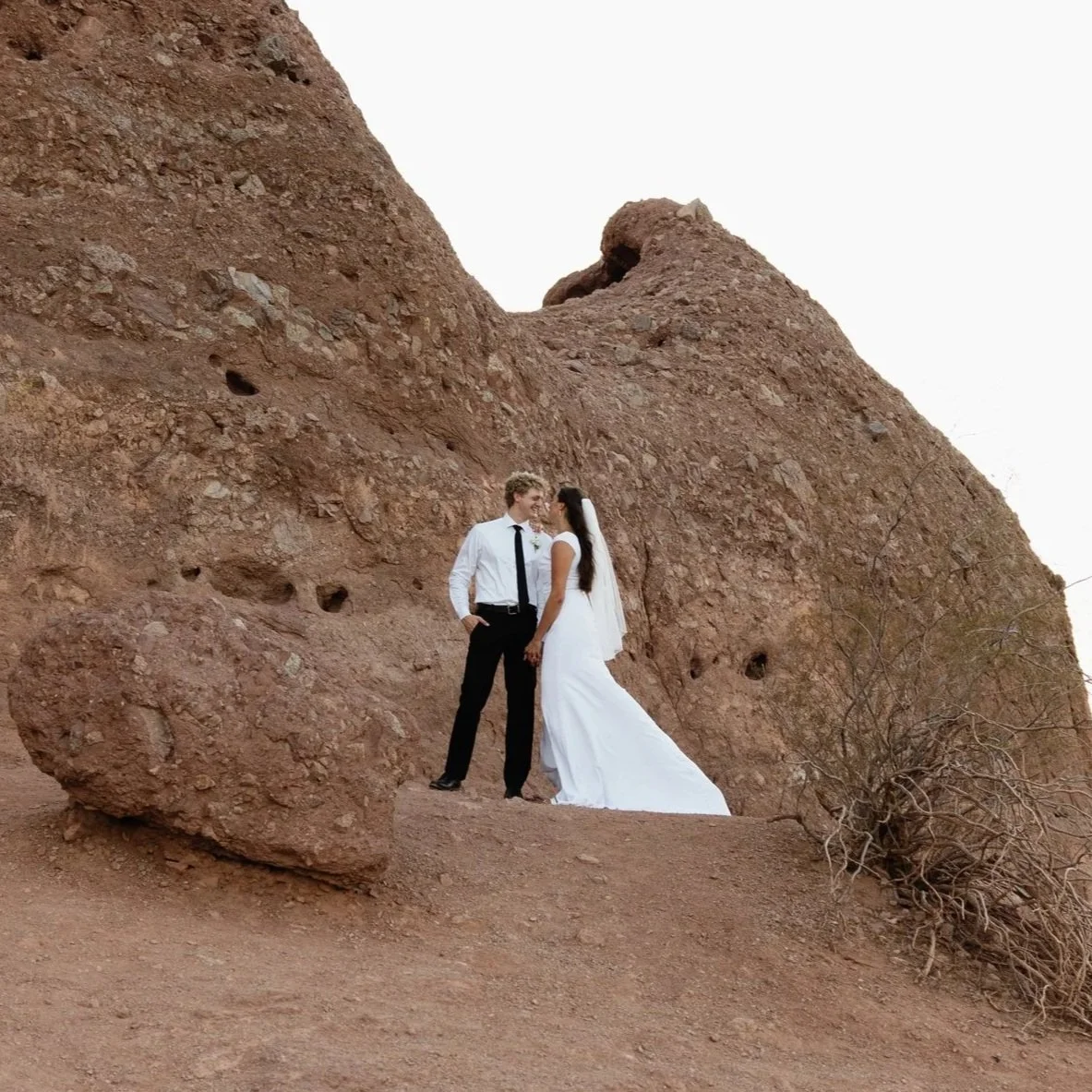 A bride and groom standing close together in front of a large reddish-brown rock formation in a desert landscape.