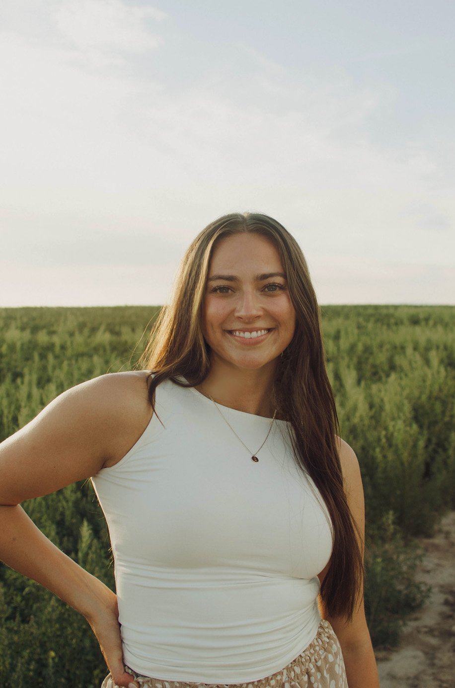 A young woman smiling outdoors in a field, wearing a white sleeveless top and necklace, with long brown hair.