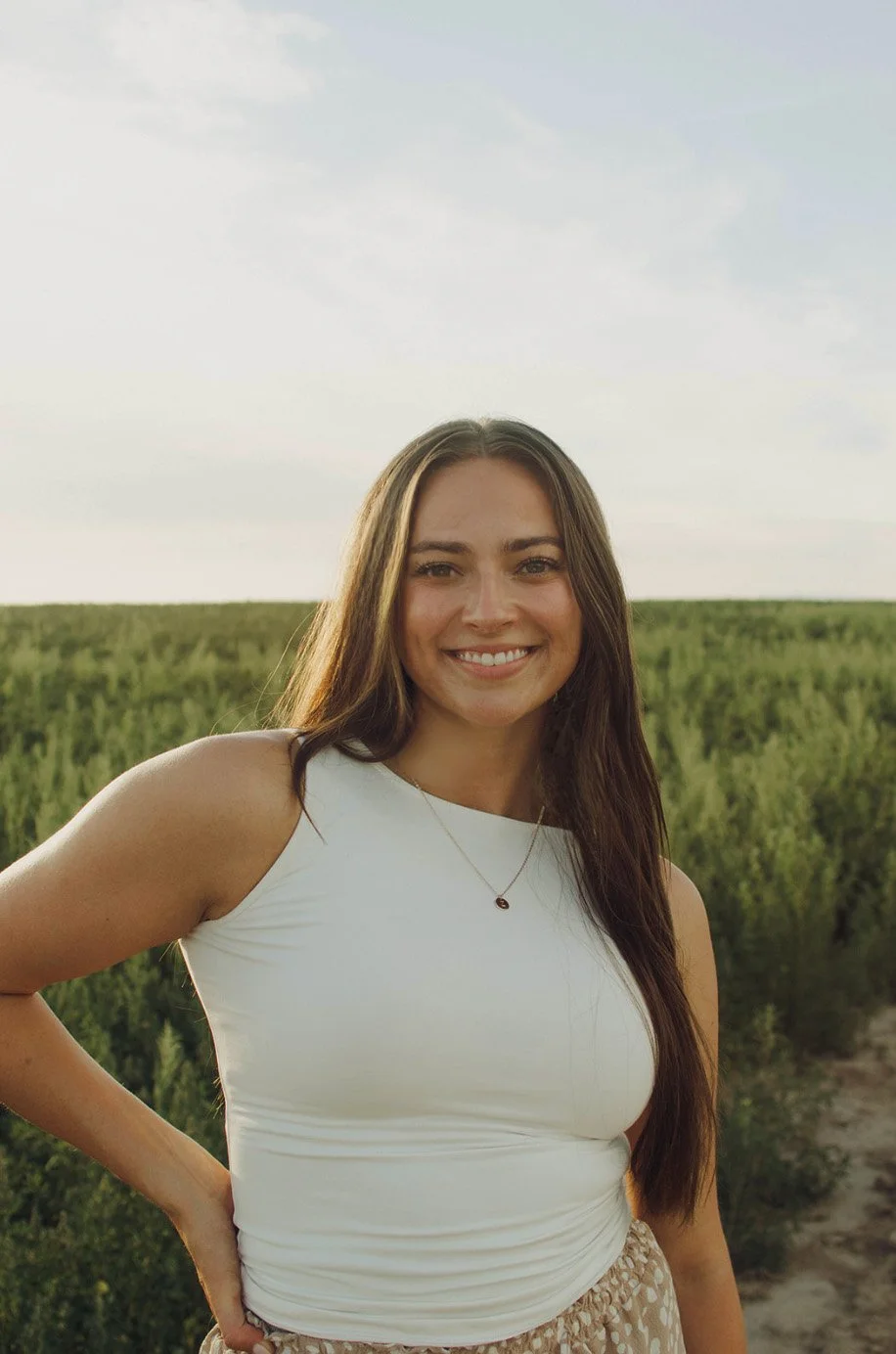 A young woman with long brown hair smiling outdoors in a green field with a partly cloudy sky.