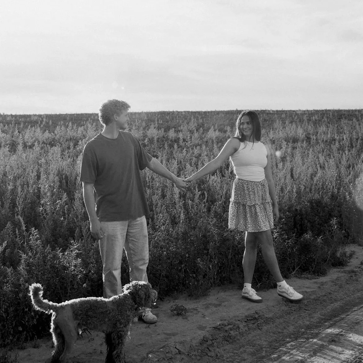 A young couple holding hands in a field with a dog walking near them during sunset in black and white.