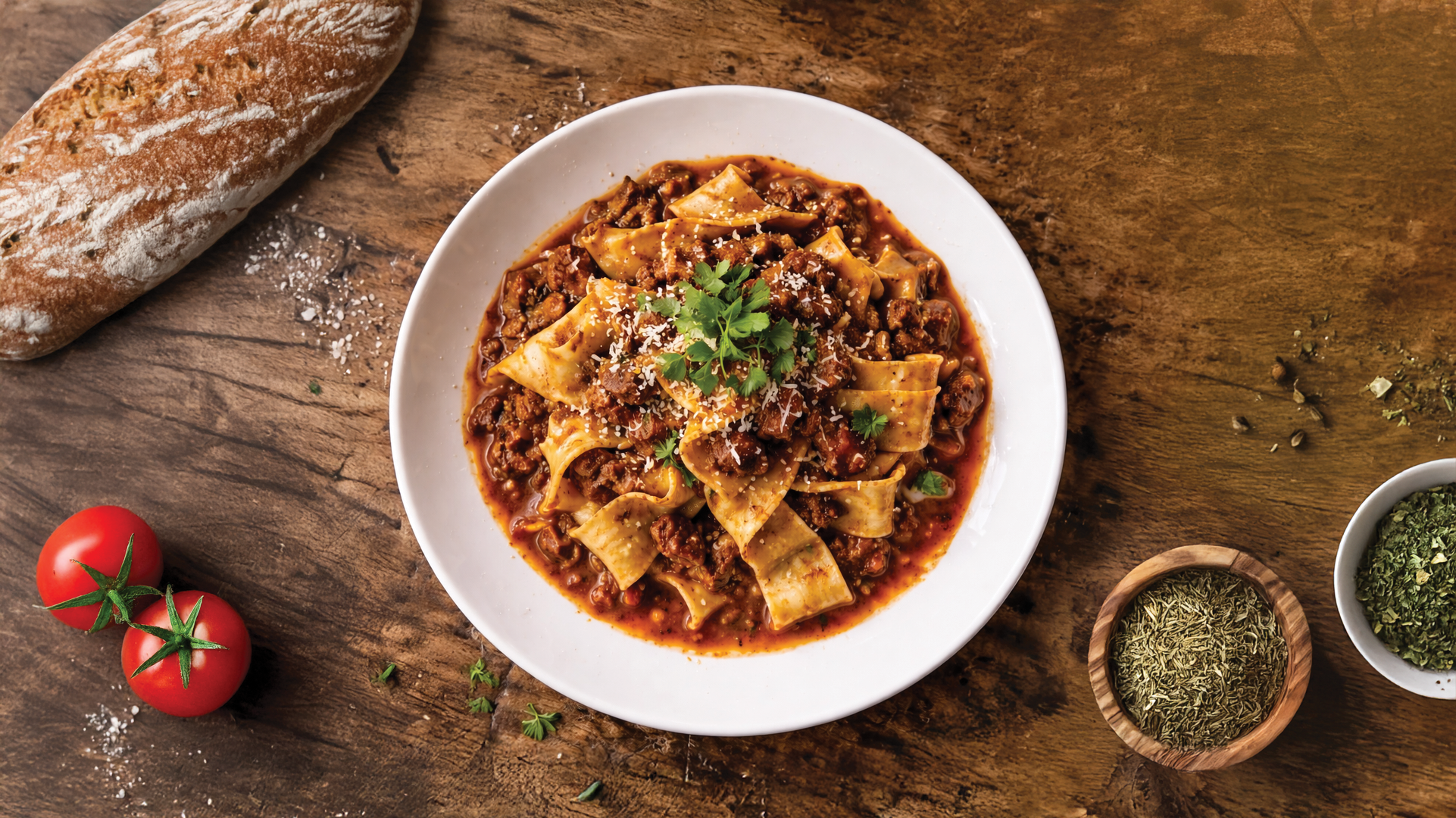 A plate of beef and pasta with tomato sauce, garnished with cilantro, on a wooden table surrounded by cherry tomatoes, a loaf of rustic bread, and small bowls of herbs and spices.