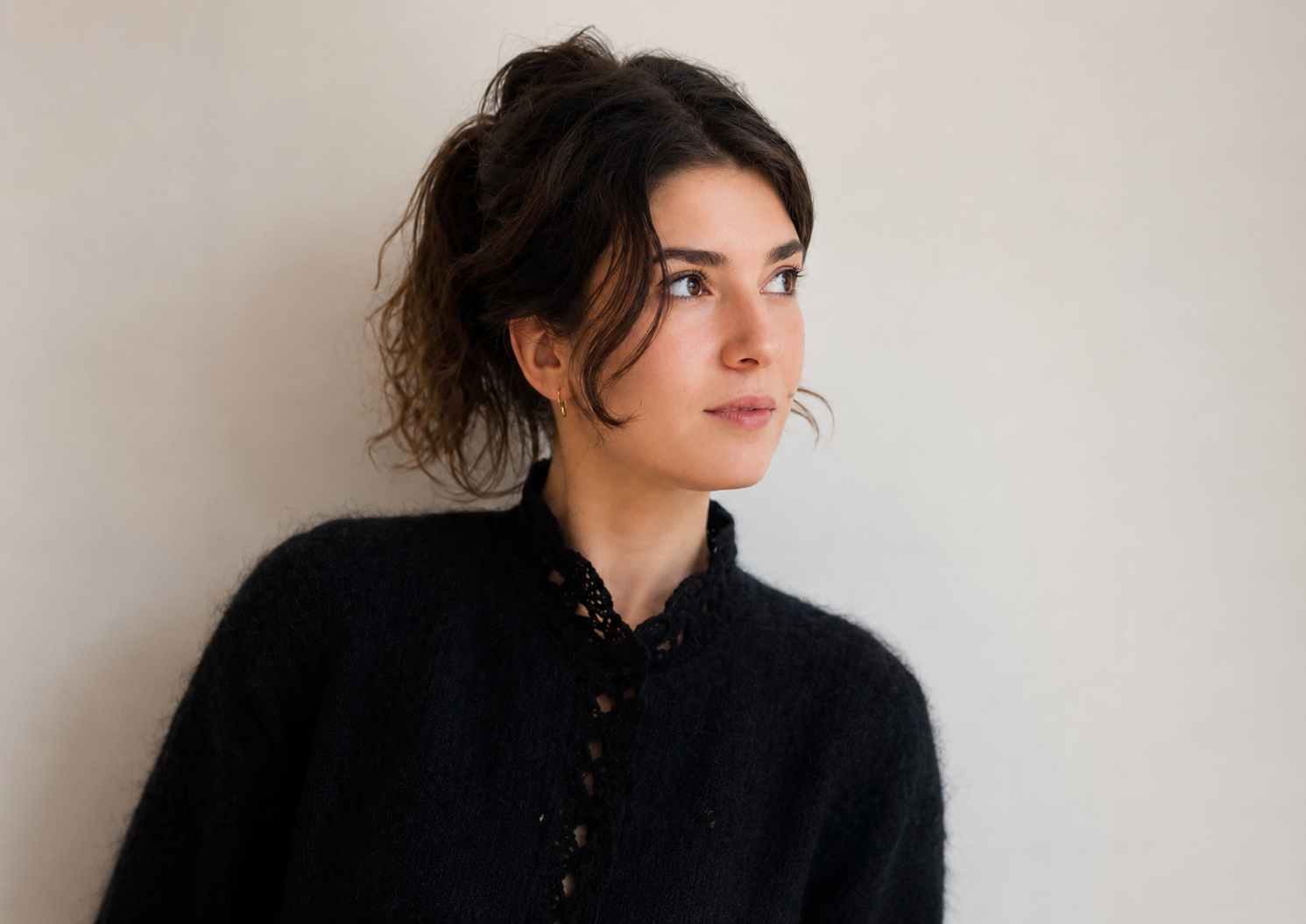 Jeune femme à cheveux bouclés et bruns, portant un pull noir avec détails en dentelle, regardant vers la droite, contre un mur beige.