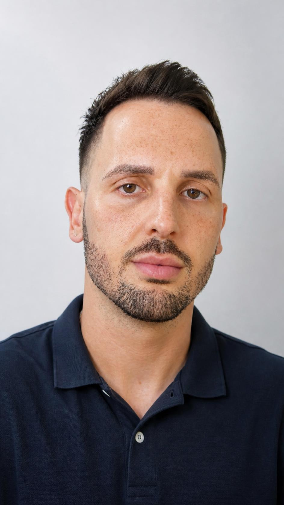 A portrait of a young man with short brown hair and facial hair, wearing a dark polo shirt, standing against a plain light gray background.