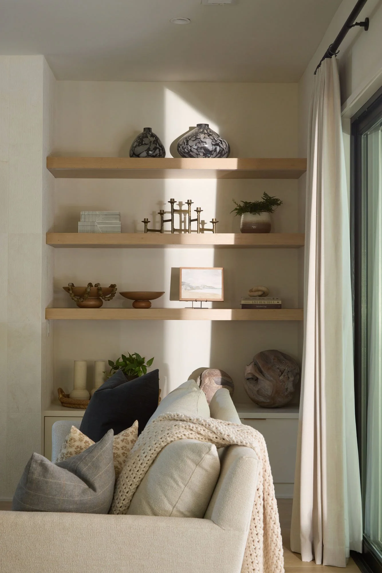 A cozy living room setting with a cream-colored sofa, decorative pillows, a knitted throw blanket, wooden shelves with decorative items like vases, candles, and plants, and a large window with white curtains.
