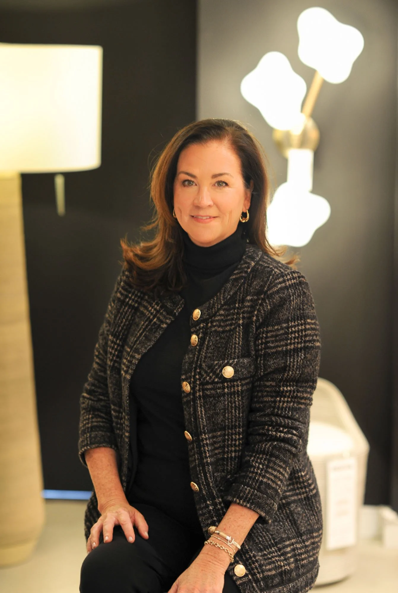 A woman with shoulder-length brown hair, wearing a black turtleneck and a black and gold plaid blazer, sitting indoors with a dark background, a lamp, and a modern decorative light in the background.