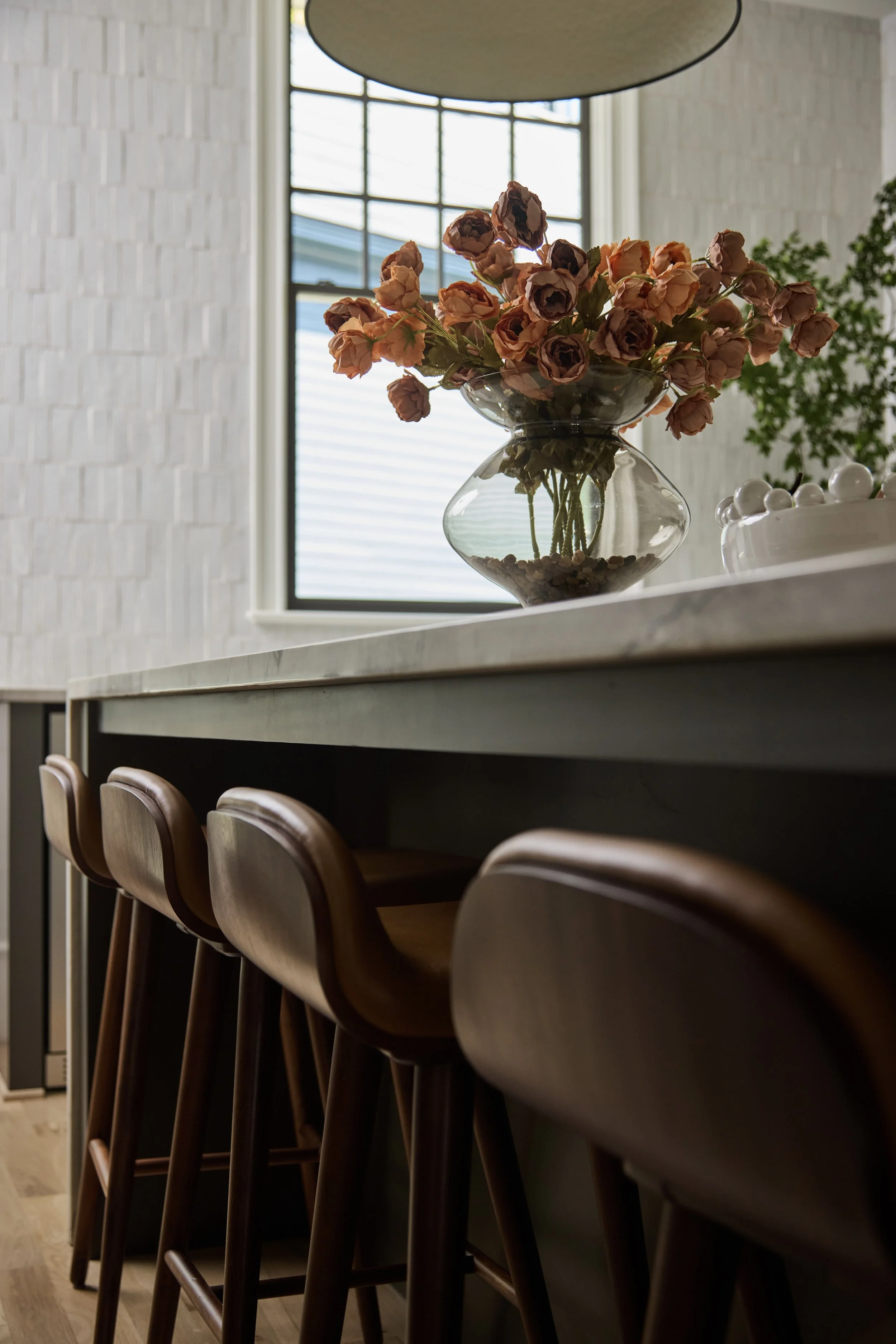 A row of brown wooden chairs aligned along a marble-topped counter in a modern interior space. There is a glass vase with pink flowers and a bowl of white decorative balls on the counter. A large window in the background lets in natural light, illuminating the textured white wall behind.