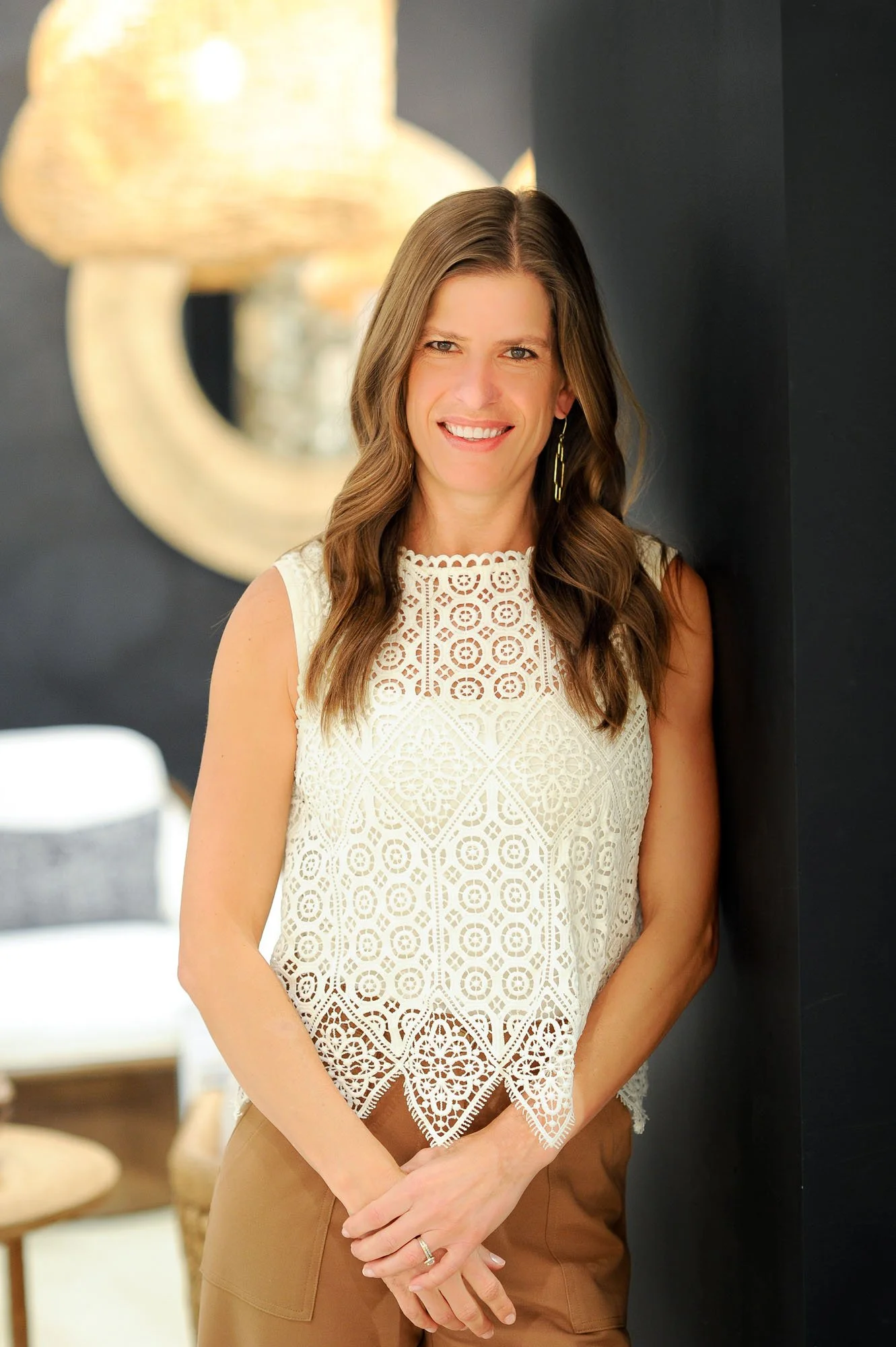 A woman with long, wavy brown hair smiling, wearing a white lace sleeveless top and tan pants, standing beside a dark wall with a large, blurry golden light fixture in the background.